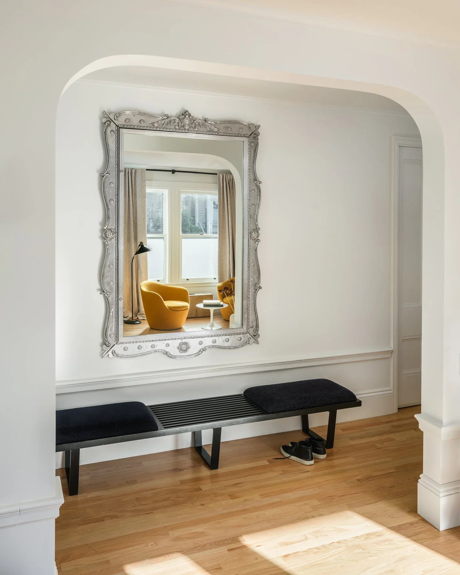 White plaster arched entryway with oversized silver ornate mirror, Nelson-style slat bench, and reflection of mustard yellow reading chairs in living room — Hill Street Residence San Francisco by Mokume Design Studio