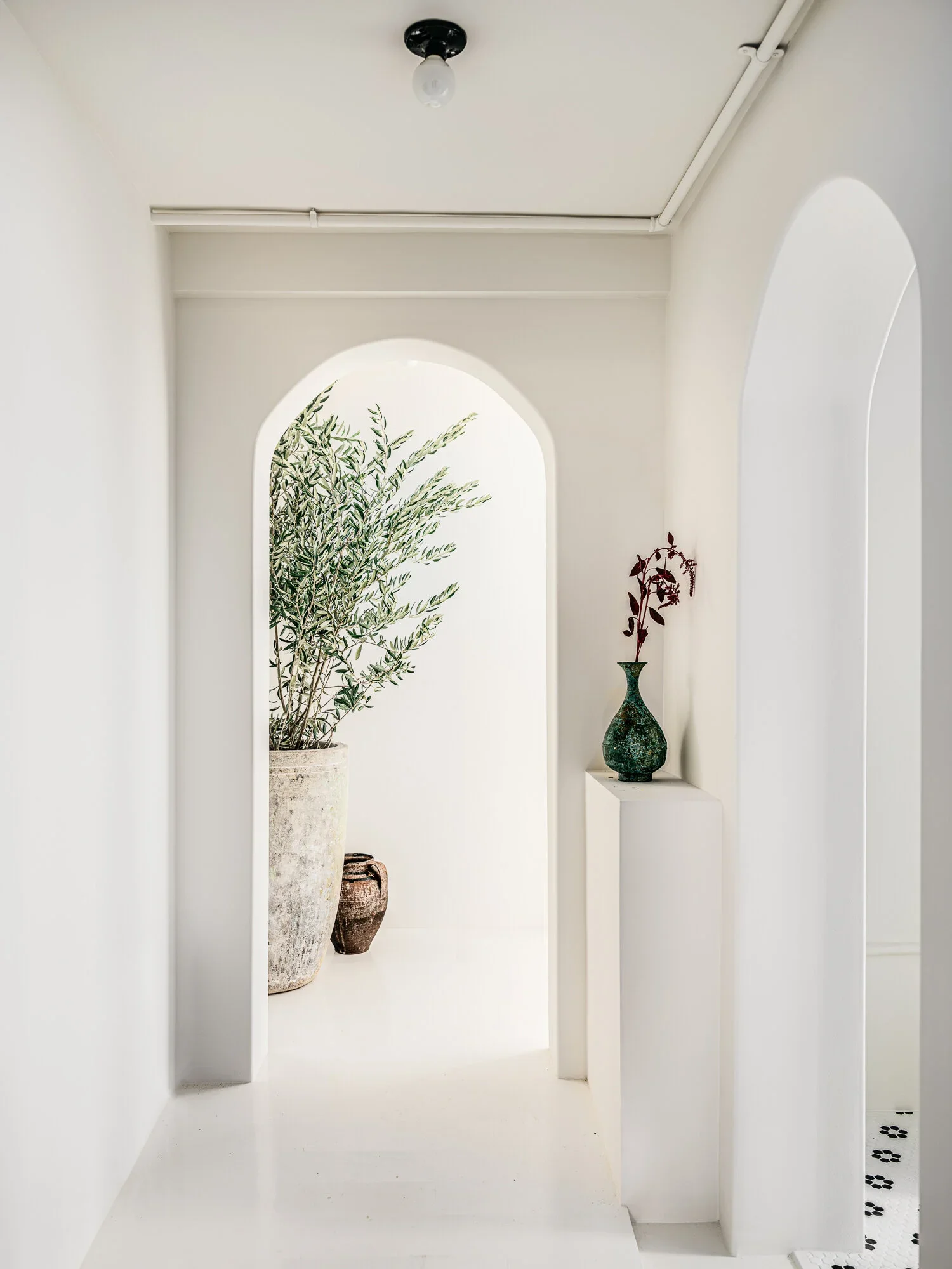 All-white arched hallway with olive tree in terracotta planter, antique ceramic vessels, and sculptural vase on plinth — Monastery Made San Francisco retail interior design by Mokume Design Studio