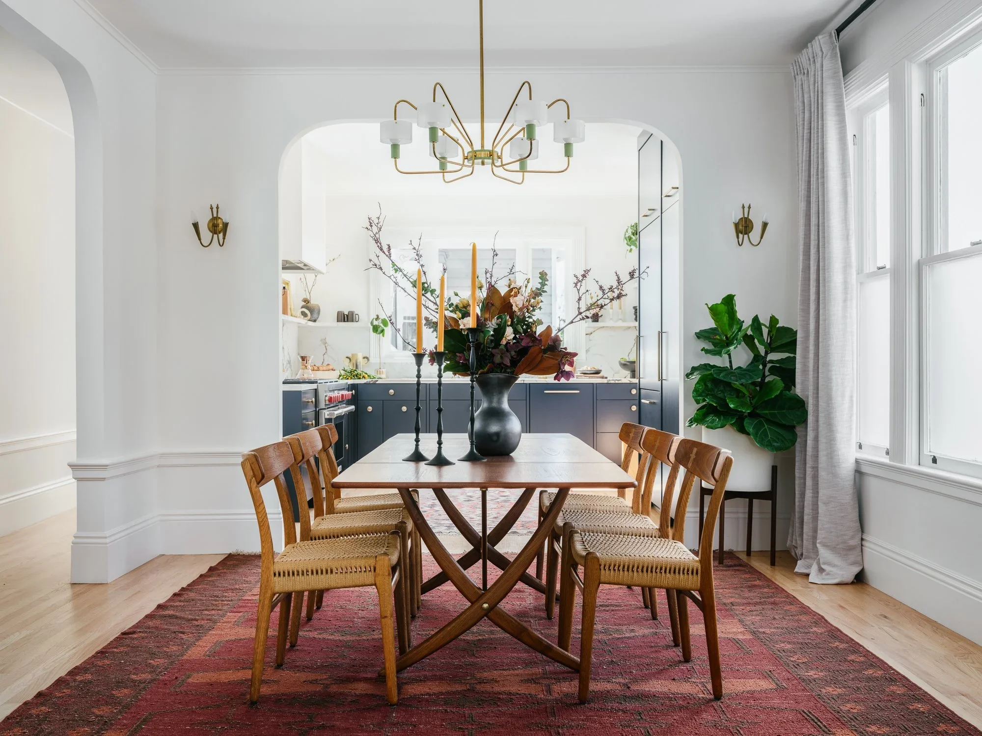 Dining room with walnut trestle table, woven seat chairs, brass chandelier, vintage red kilim rug, and arched opening to navy blue kitchen beyond — Hill Street Residence San Francisco by Mokume Design Studio