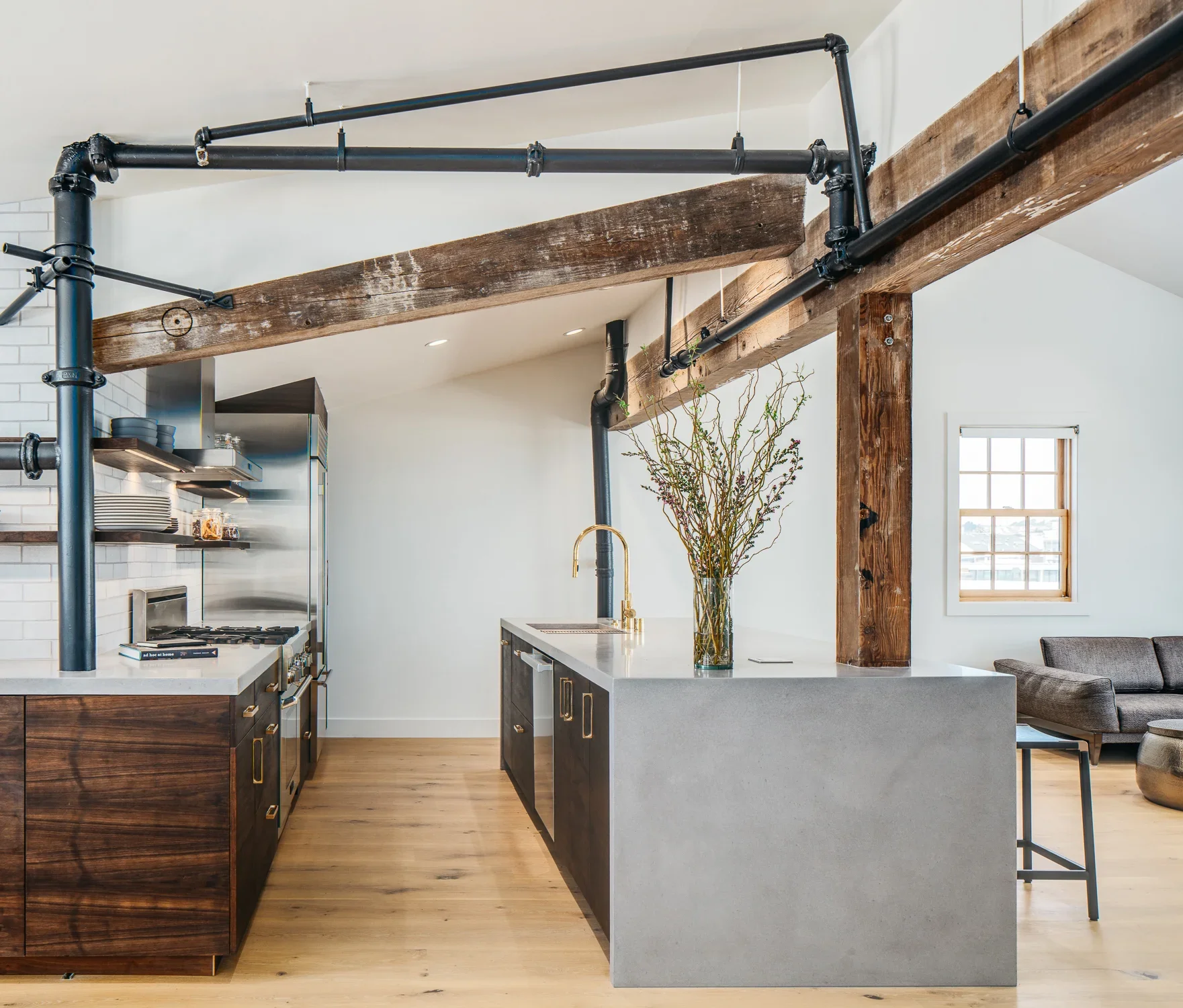 Open-concept industrial loft kitchen with exposed black iron pipe, reclaimed wood ceiling beams, dark walnut cabinetry, concrete waterfall island, and adjacent living area — Mission Loft San Francisco by Mokume Design Studio