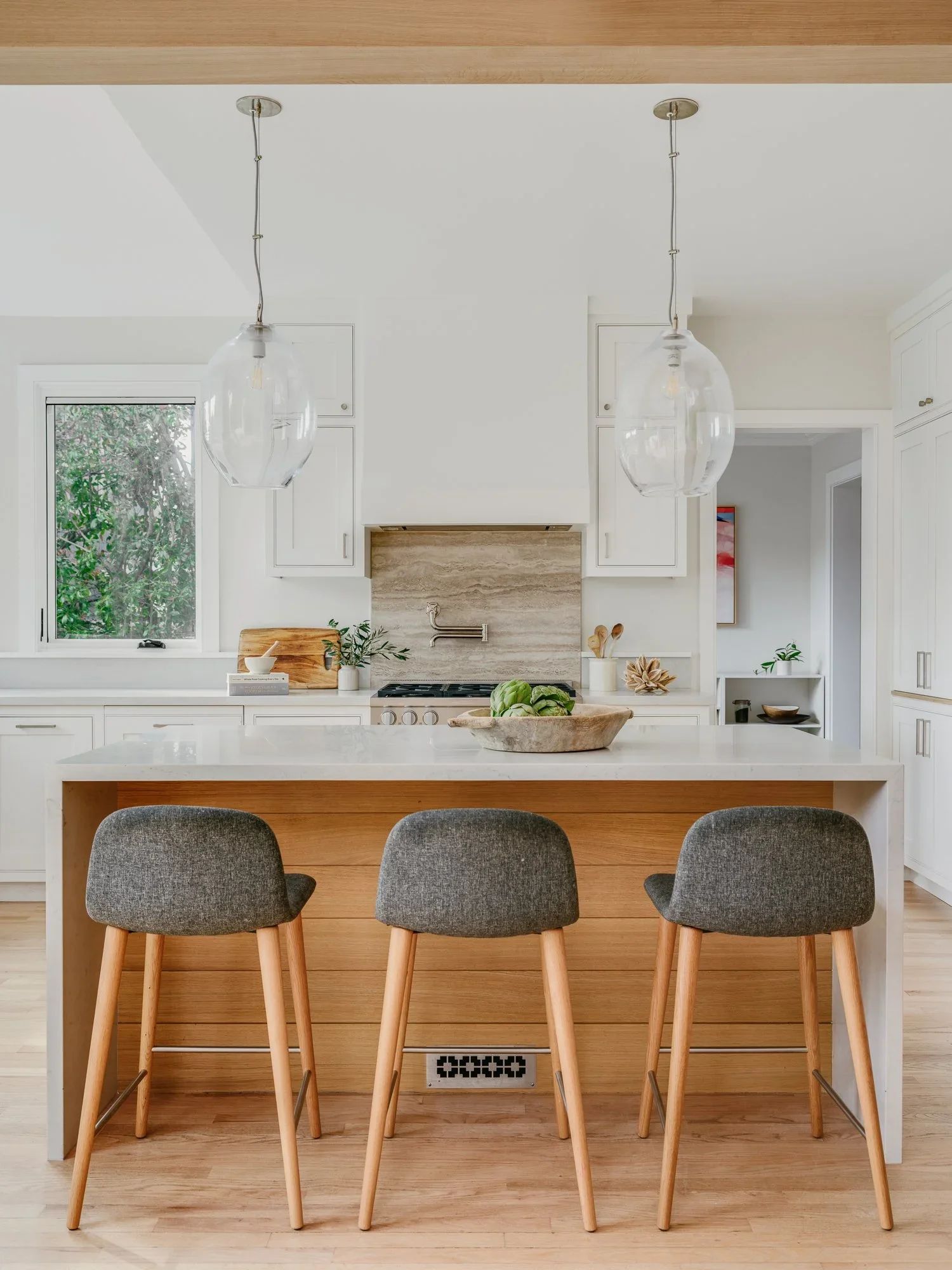 Bright open-concept kitchen with white quartz island, oak beam ceiling, globe glass pendants, farmhouse sink bay window, and travertine range backsplash — Kentfield California home remodel by Mokume Design Studio