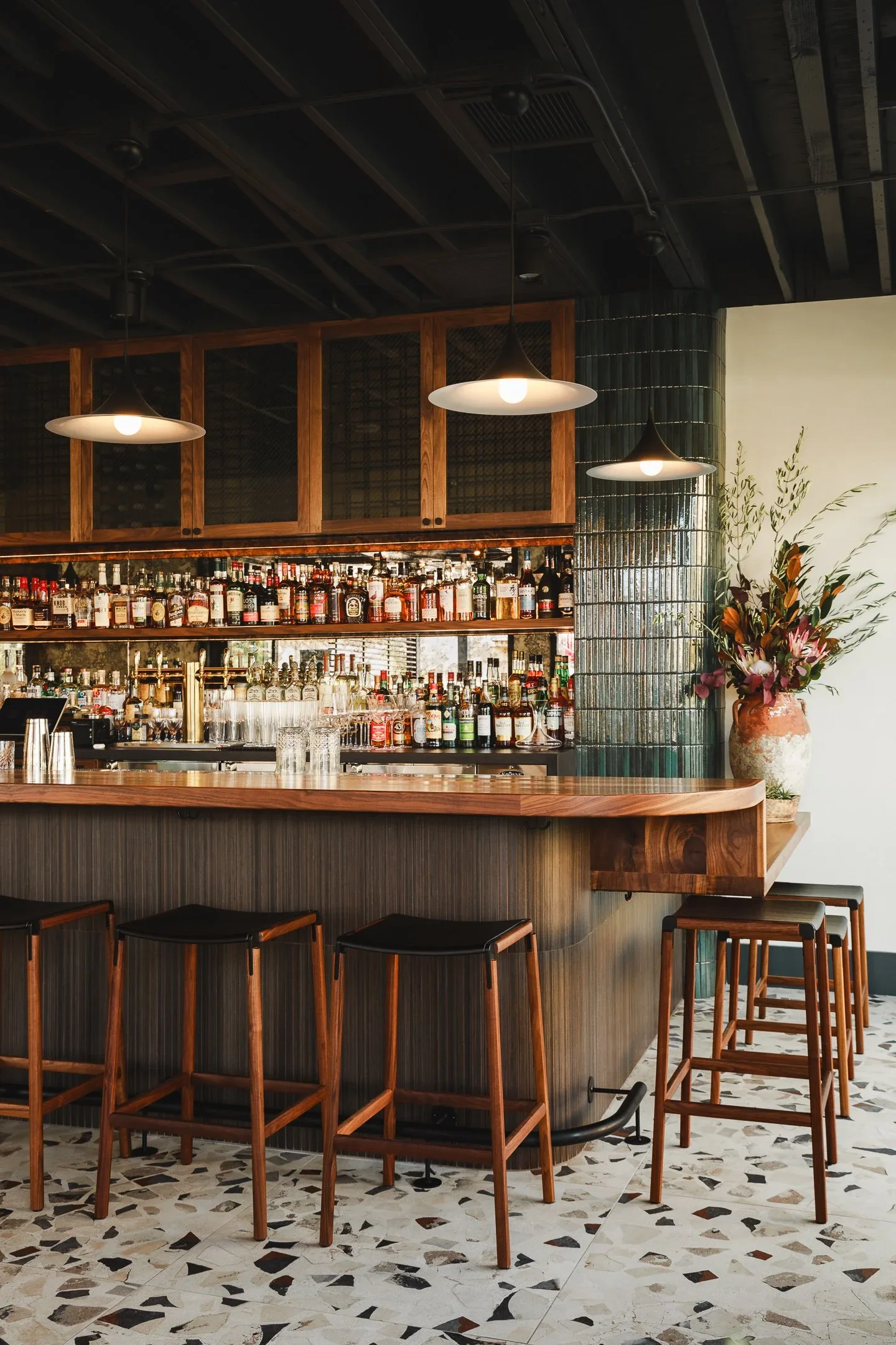 Lively restaurant dining room with set tables, amber candlelight, and glowing full-service bar visible in the background — Marlena restaurant Long Beach by Mokume Design Studio