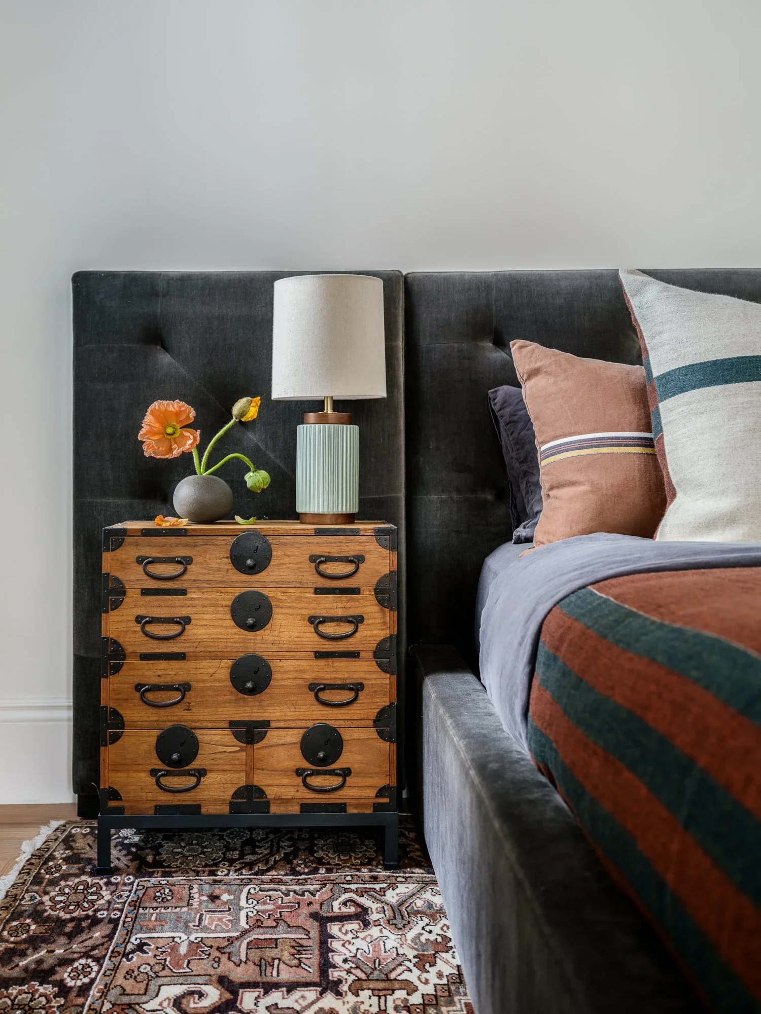 Close-up of antique Japanese tansu chest nightstand with ceramic table lamp, black ceramic bud vase, and orange poppy florals beside charcoal velvet bed — Hill Street Residence San Francisco by Mokume Design Studio