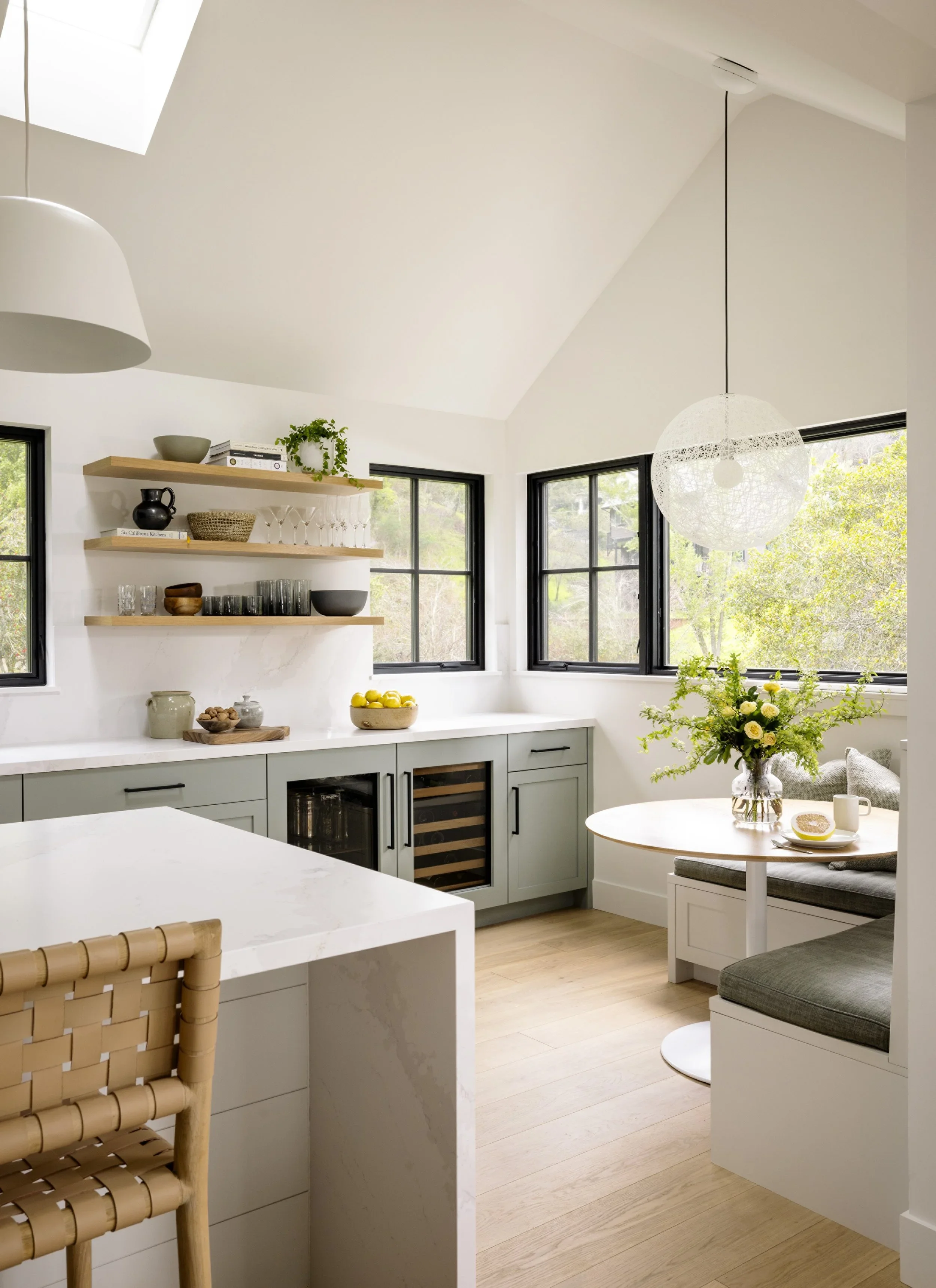Sage green shaker kitchen with white quartz island, woven leather barstools, open oak shelving, wine fridge, and breakfast nook beyond — Corte Madera Residence by Mokume Design Studio