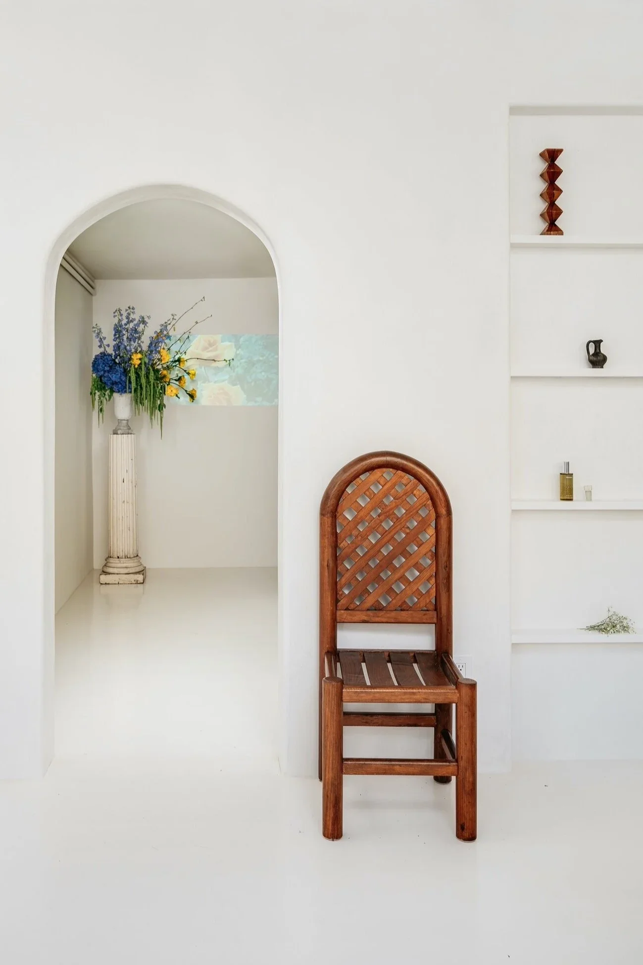 White plaster arch framing a floral installation on a classical column pedestal, with antique lattice-back wood chair and built-in display shelving — Monastery Made San Francisco retail interior by Mokume Design Studio
