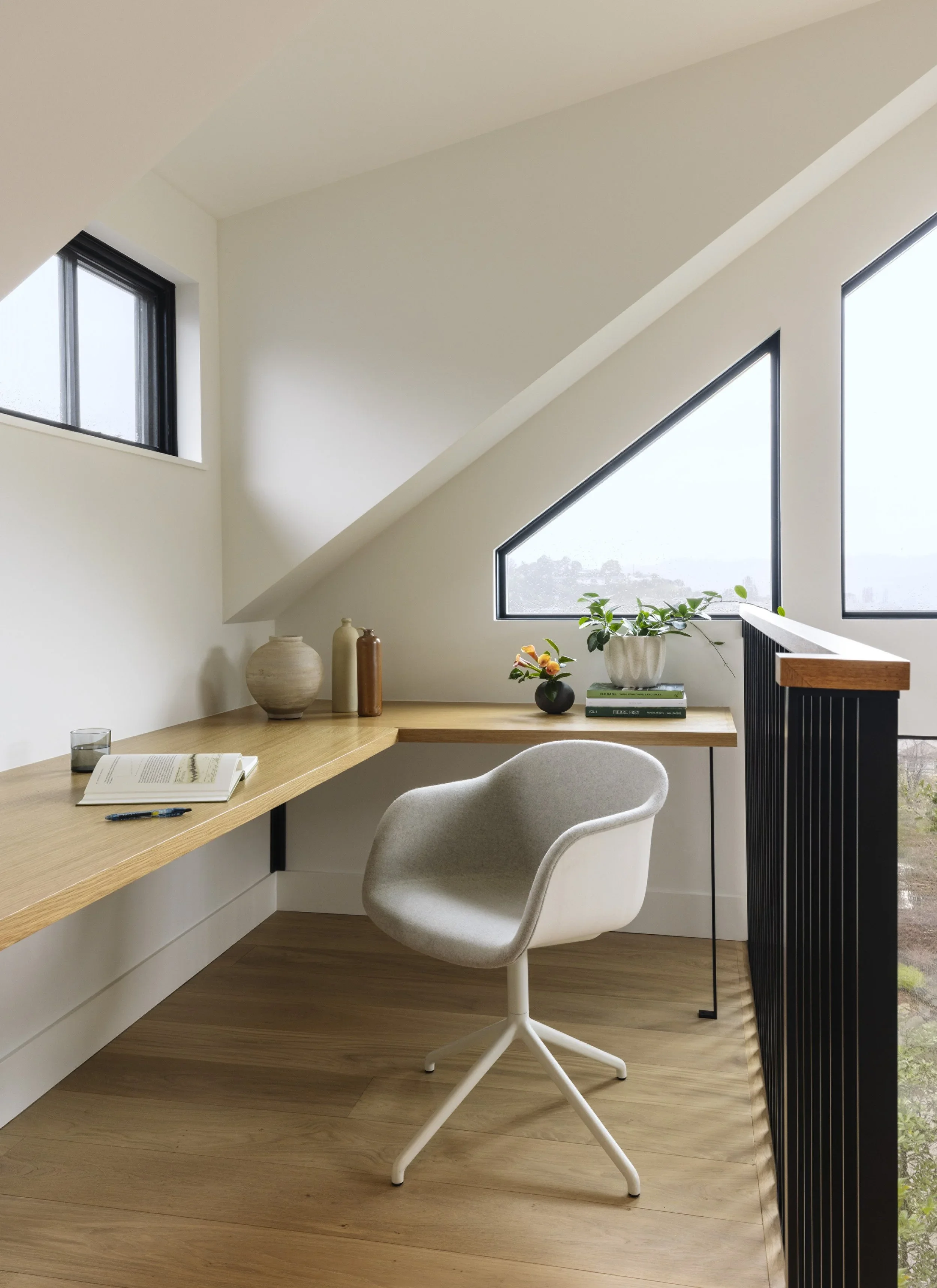 Floating natural oak L-shaped desk built into a bright loft space with angled black-framed windows, gray swivel chair, and black slatted stair railing — Corte Madera Residence by Mokume Design Studio