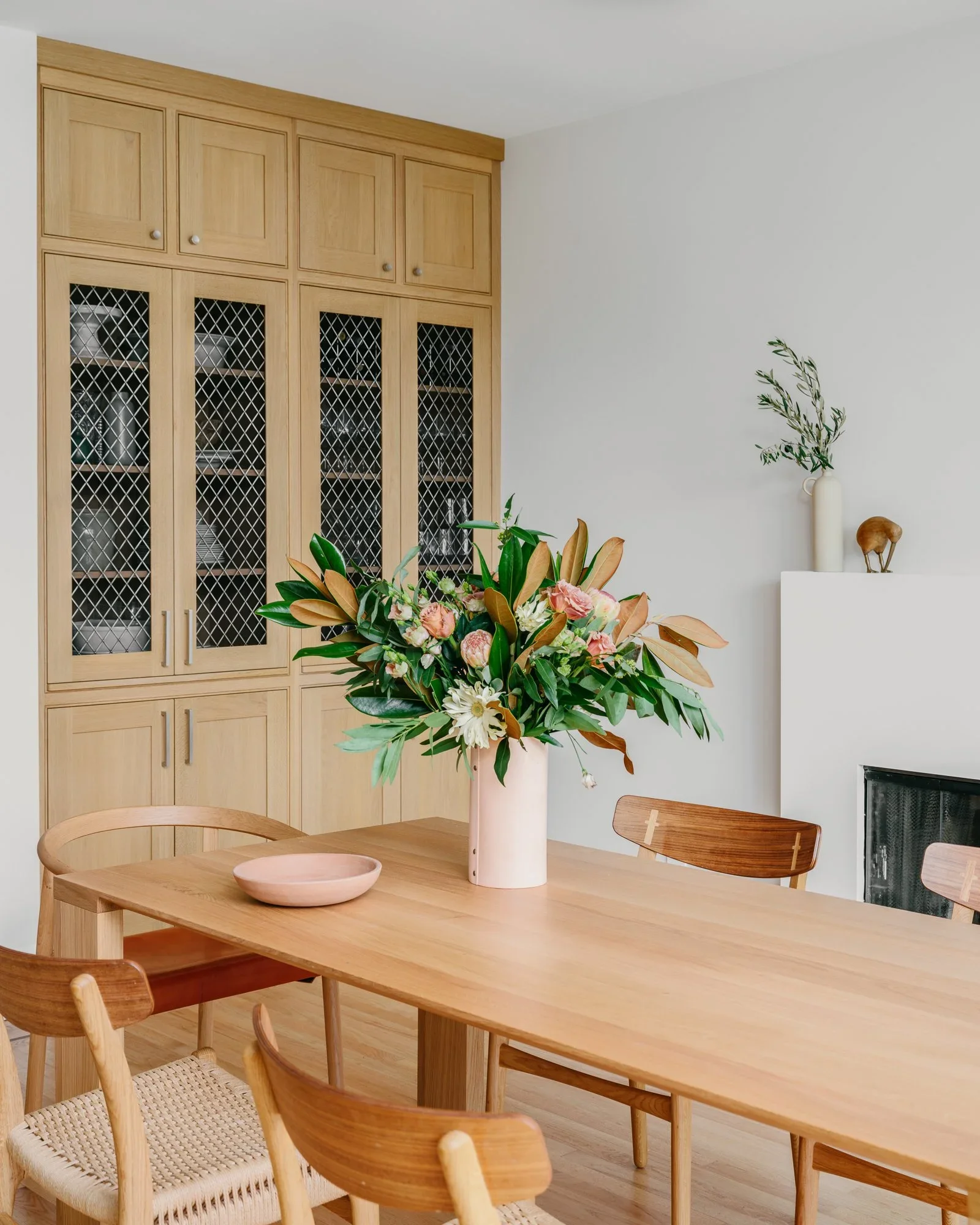 Natural oak dining room with built-in hutch, wire mesh cabinet doors, oval dining table, mixed wood chairs, and fireplace — Kentfield California home remodel by Mokume Design Studio