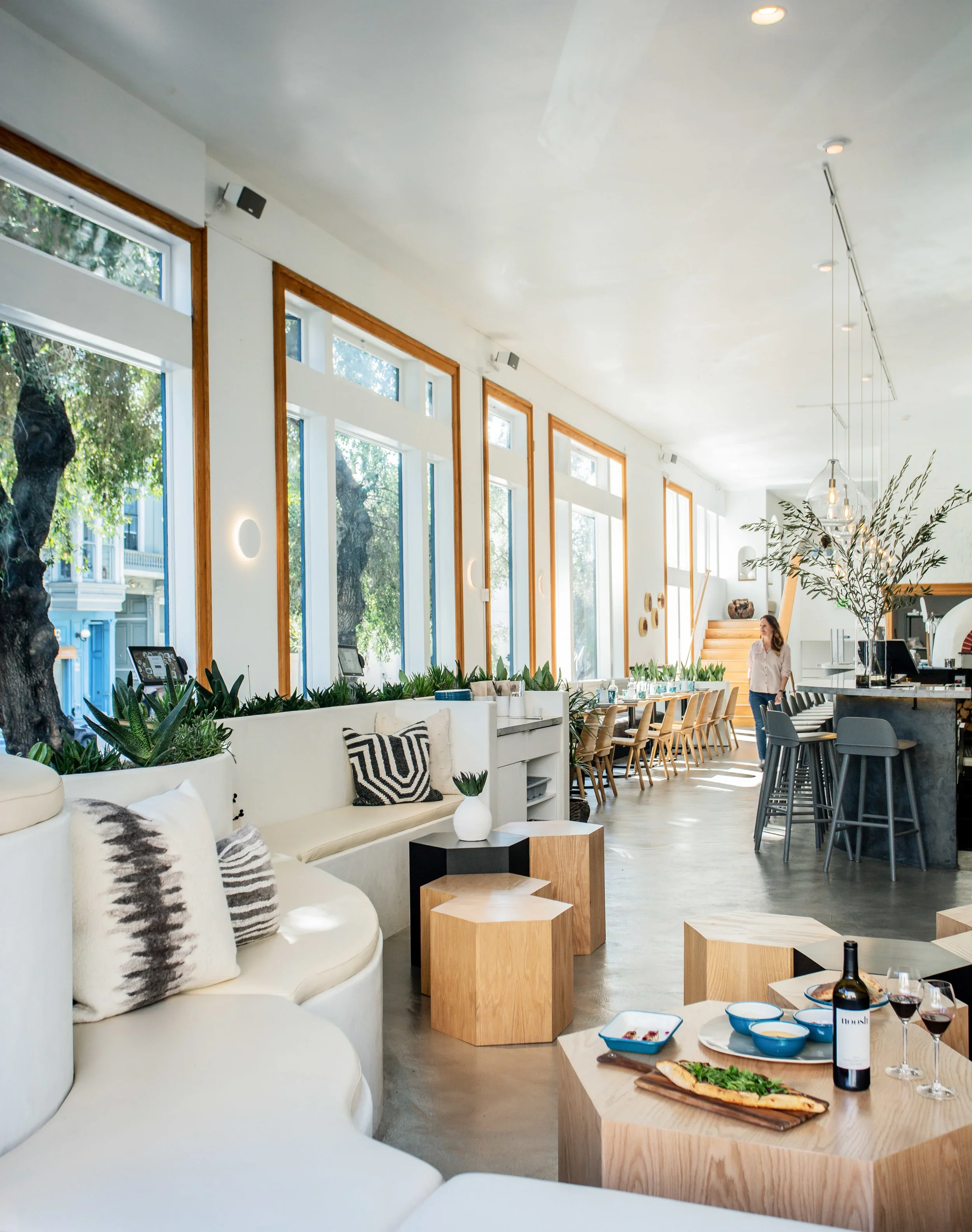 Bright open dining room at Noosh restaurant with curved white banquette, hexagonal oak side tables, floor-to-ceiling wood-framed windows, and concrete bar beyond — San Francisco restaurant design by Mokume Design Studio