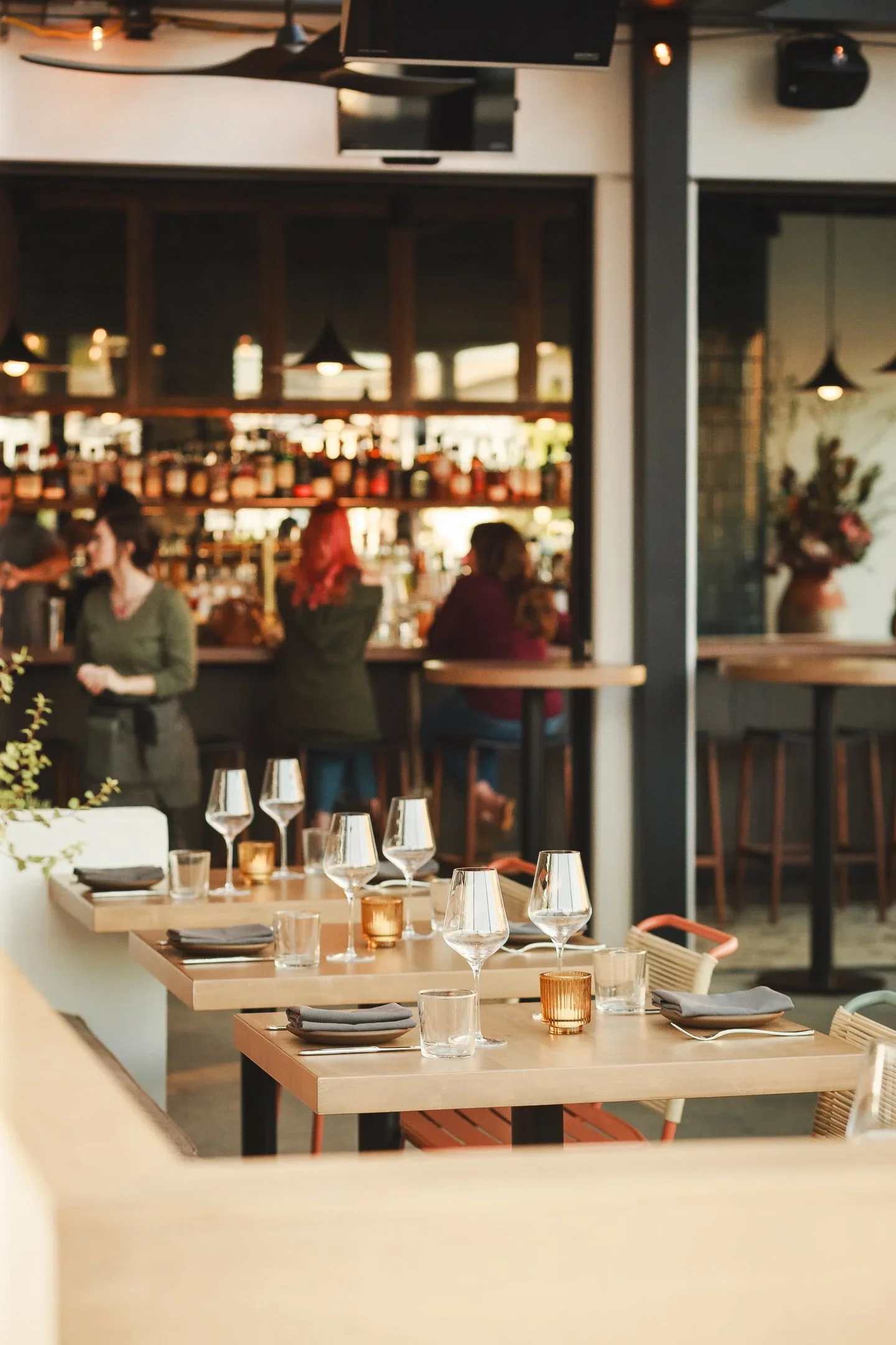 Sunlit outdoor dining table set with crystal wine glasses and amber glass votive candle against teal slatted privacy screen — Marlena restaurant Long Beach by Mokume Design Studio