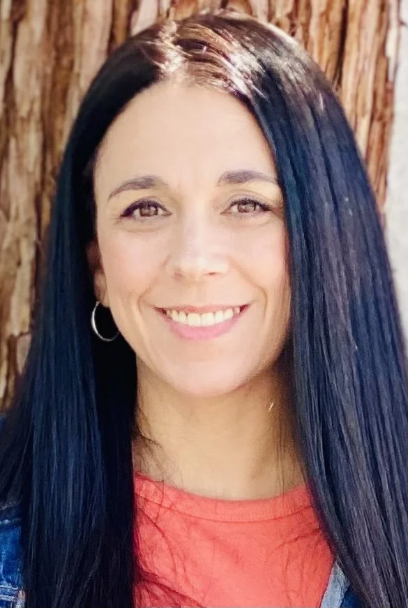A smiling woman with long black hair and hoop earrings, standing outdoors against a wooden background.