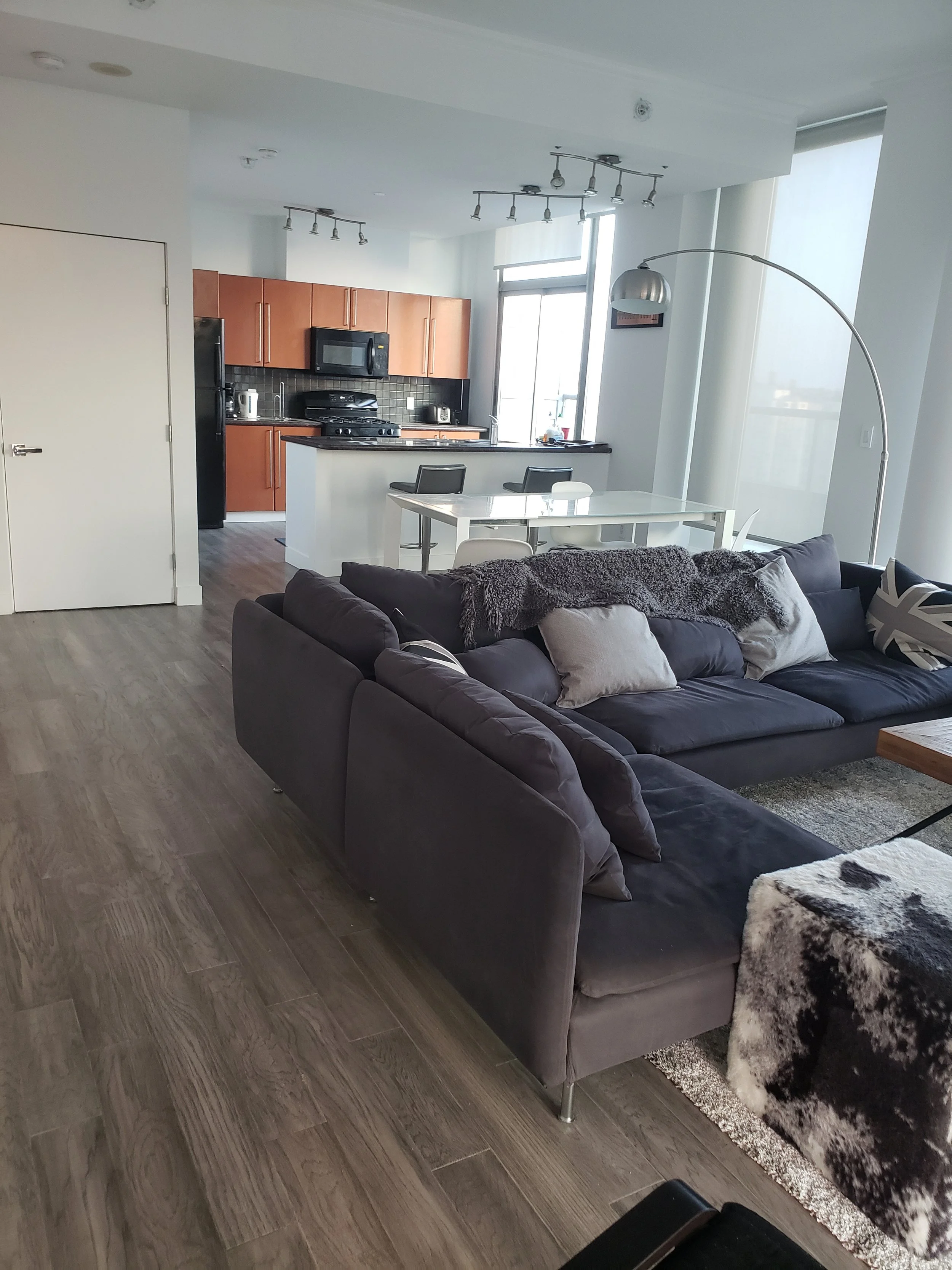 Modern open-concept apartment living room with dark gray sectional sofa, white cushions, and a cozy blanket. Adjacent to the living area is a dining table with chairs, and the kitchen with wooden cabinets, black appliances, and a breakfast bar.
