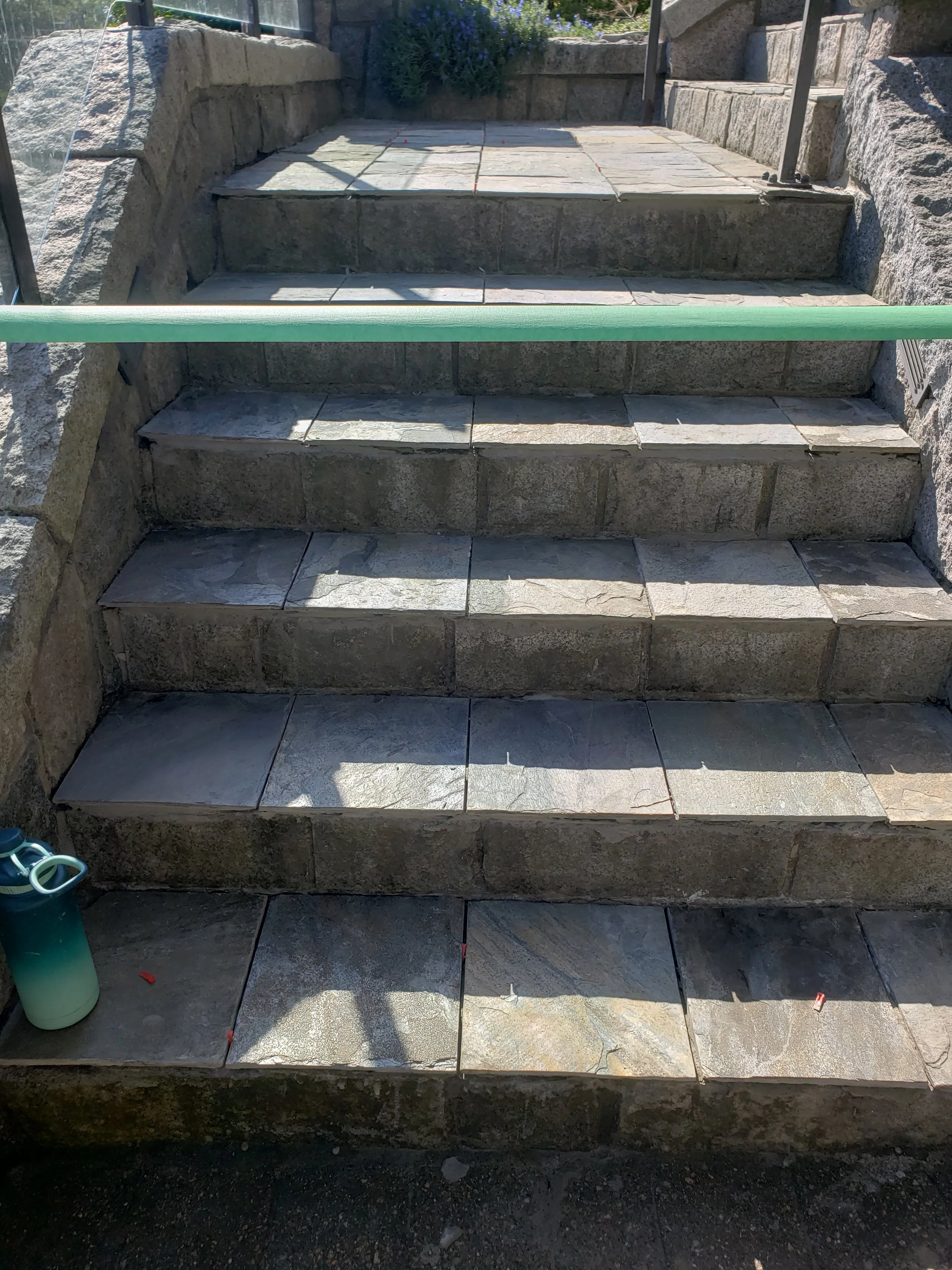 Outdoor stone staircase with four steps, bordered by stone walls on either side, and a green handrail on top of the middle wall. There is a pitcher with a small container on the bottom step. Shadows cast across the steps.