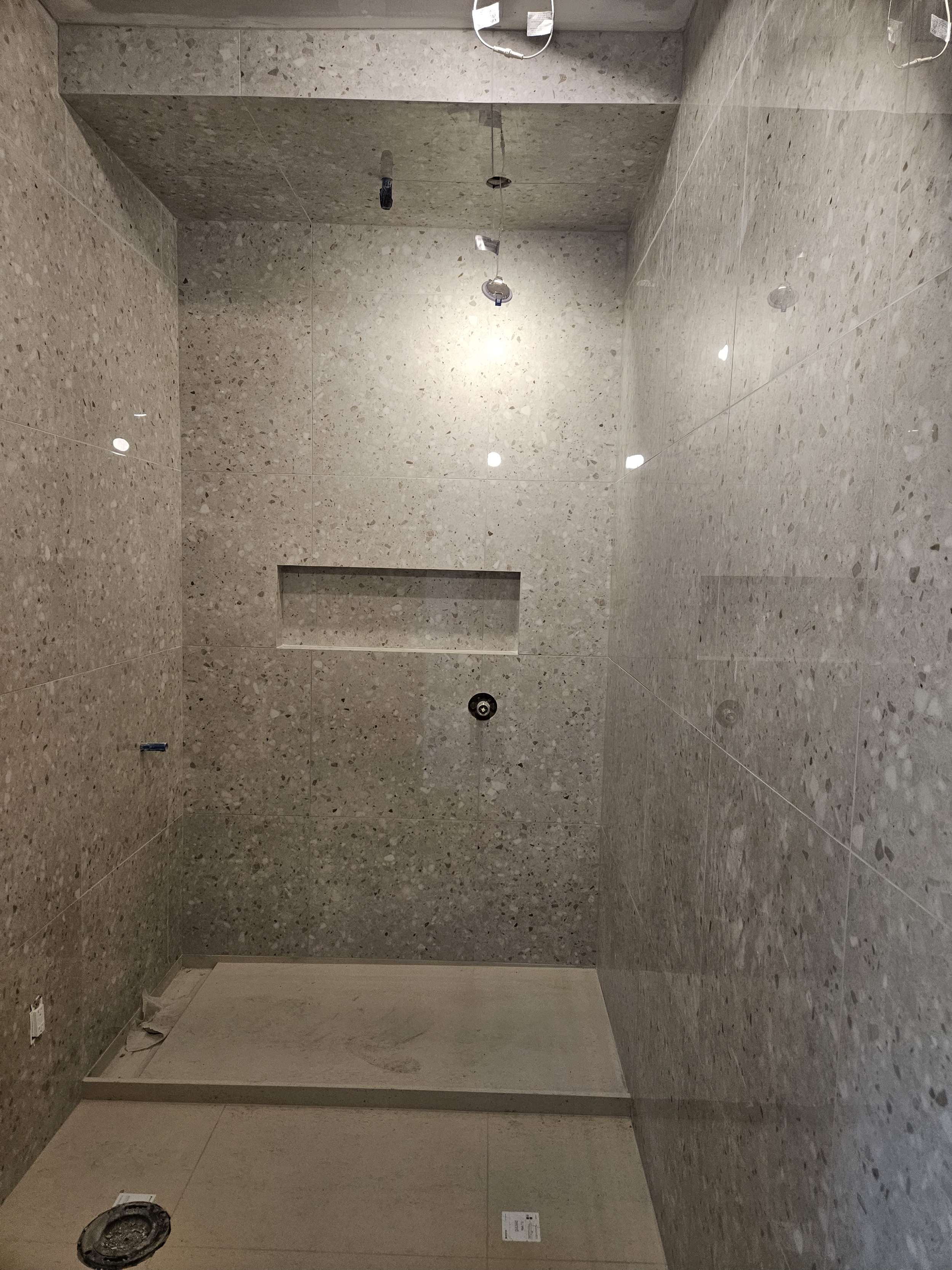 Empty shower stall with gray tiled walls, ledge, and plumbing fixtures, under construction.