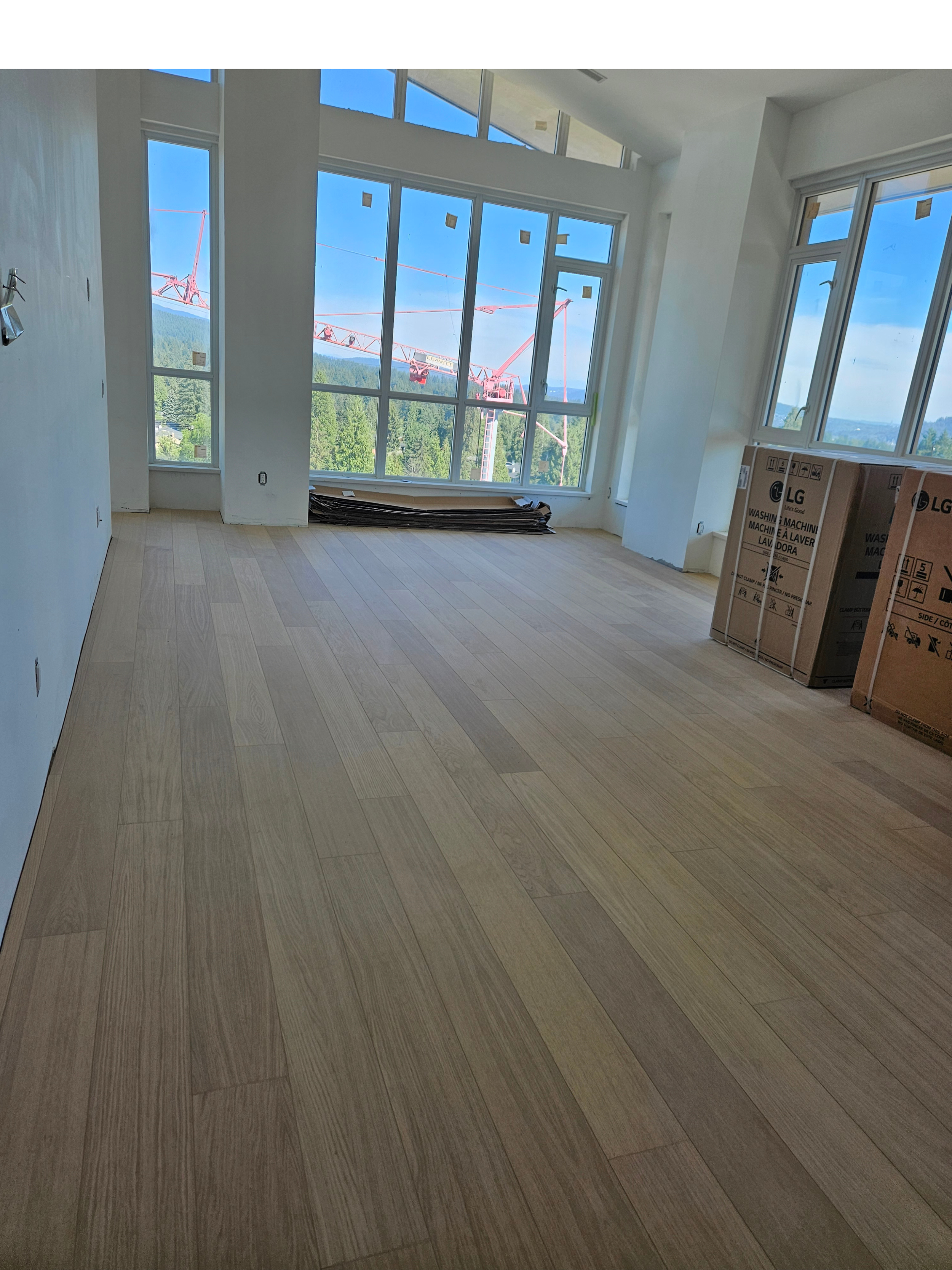 An interior room under construction with light wood flooring and large windows providing a view of construction cranes outside.