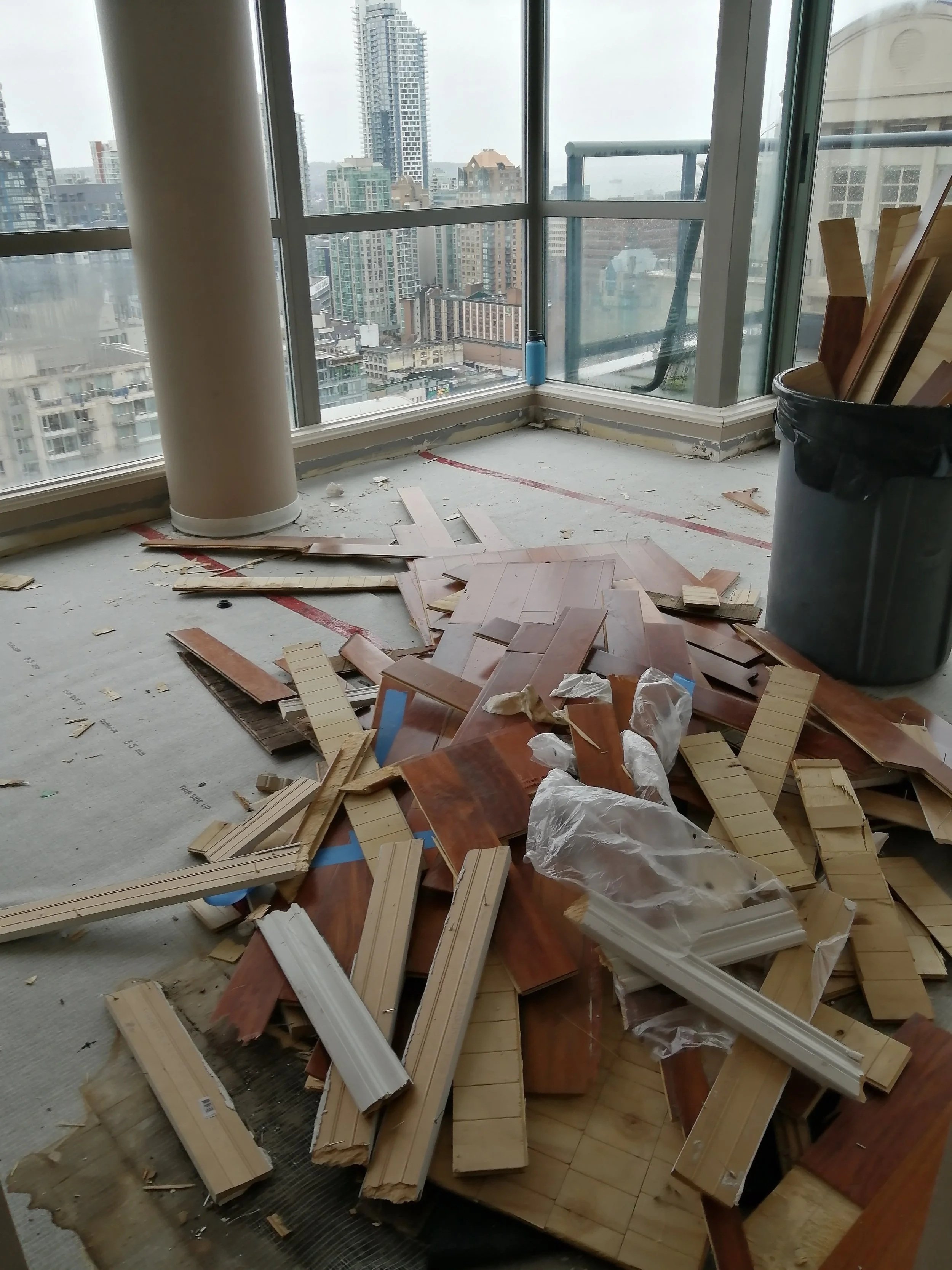 Room with scattered broken wooden flooring pieces and debris on the floor, large glass windows with a city skyline view, and construction materials and trash around.
