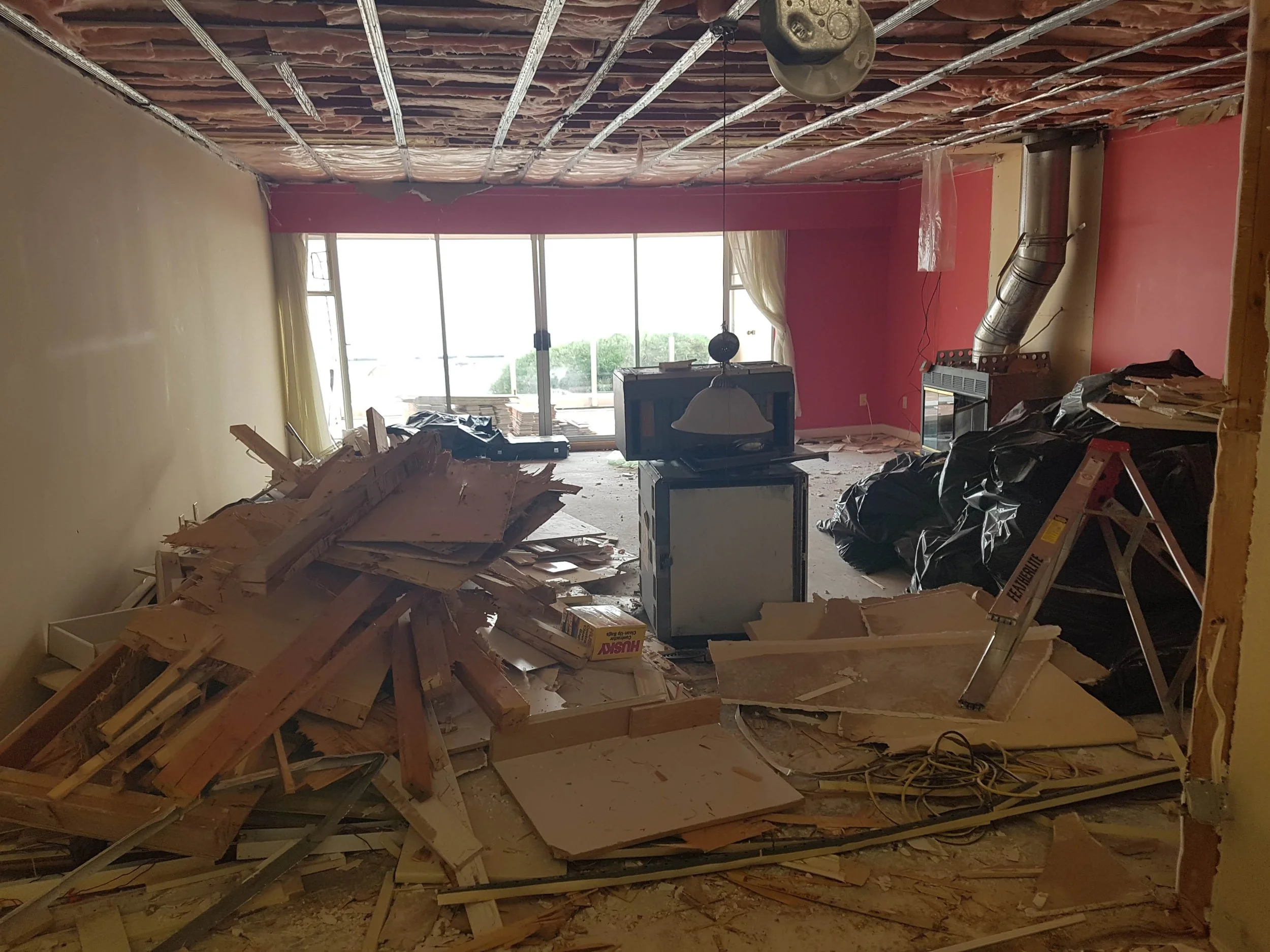 Room under renovation with construction debris and materials scattered on the floor, a ladder, a small television, black garbage bags, and a large window with curtains.