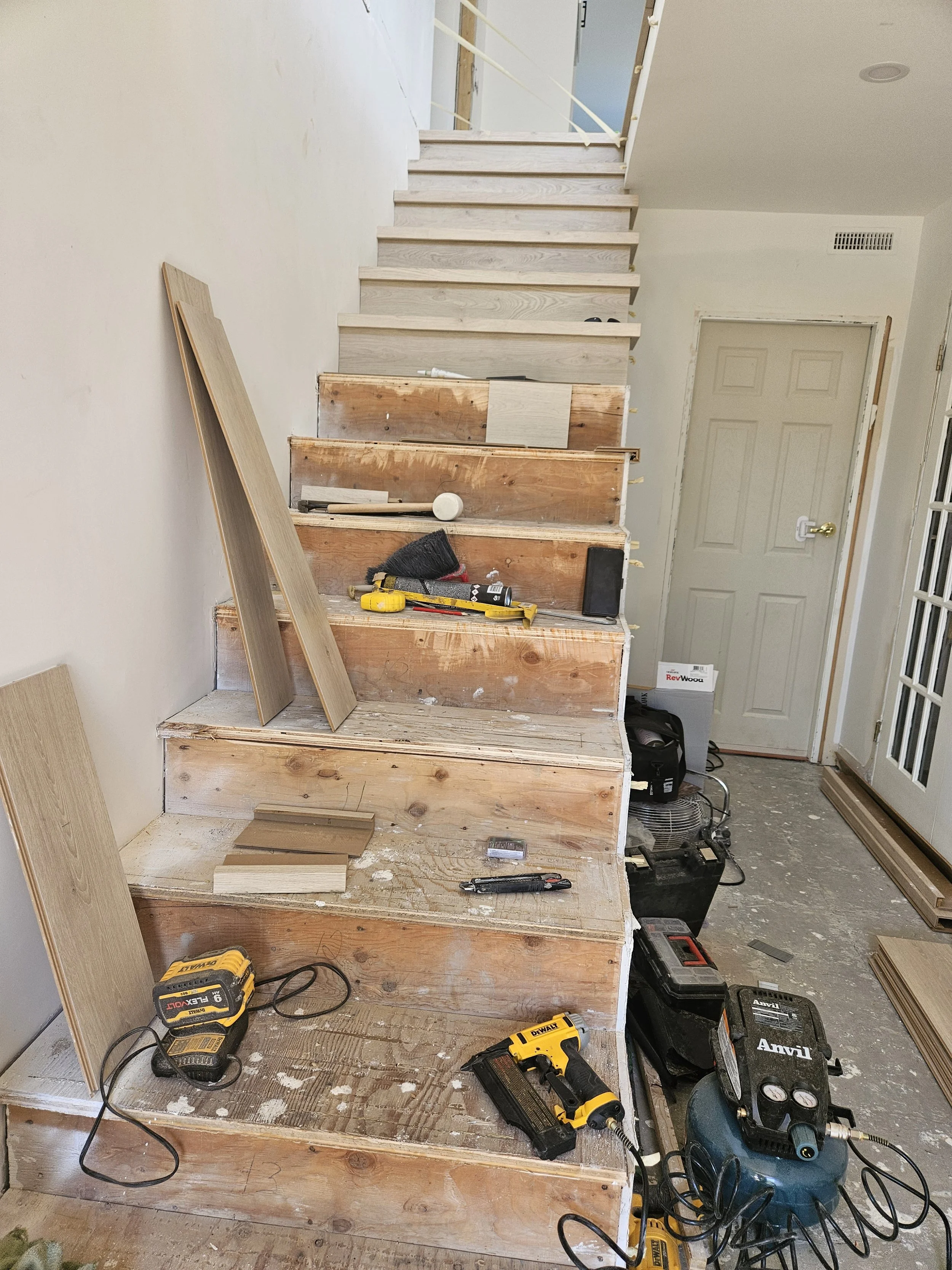 Staircase under construction with tools and materials, including drills, wood panels, and a carpet brush, in a house interior.
