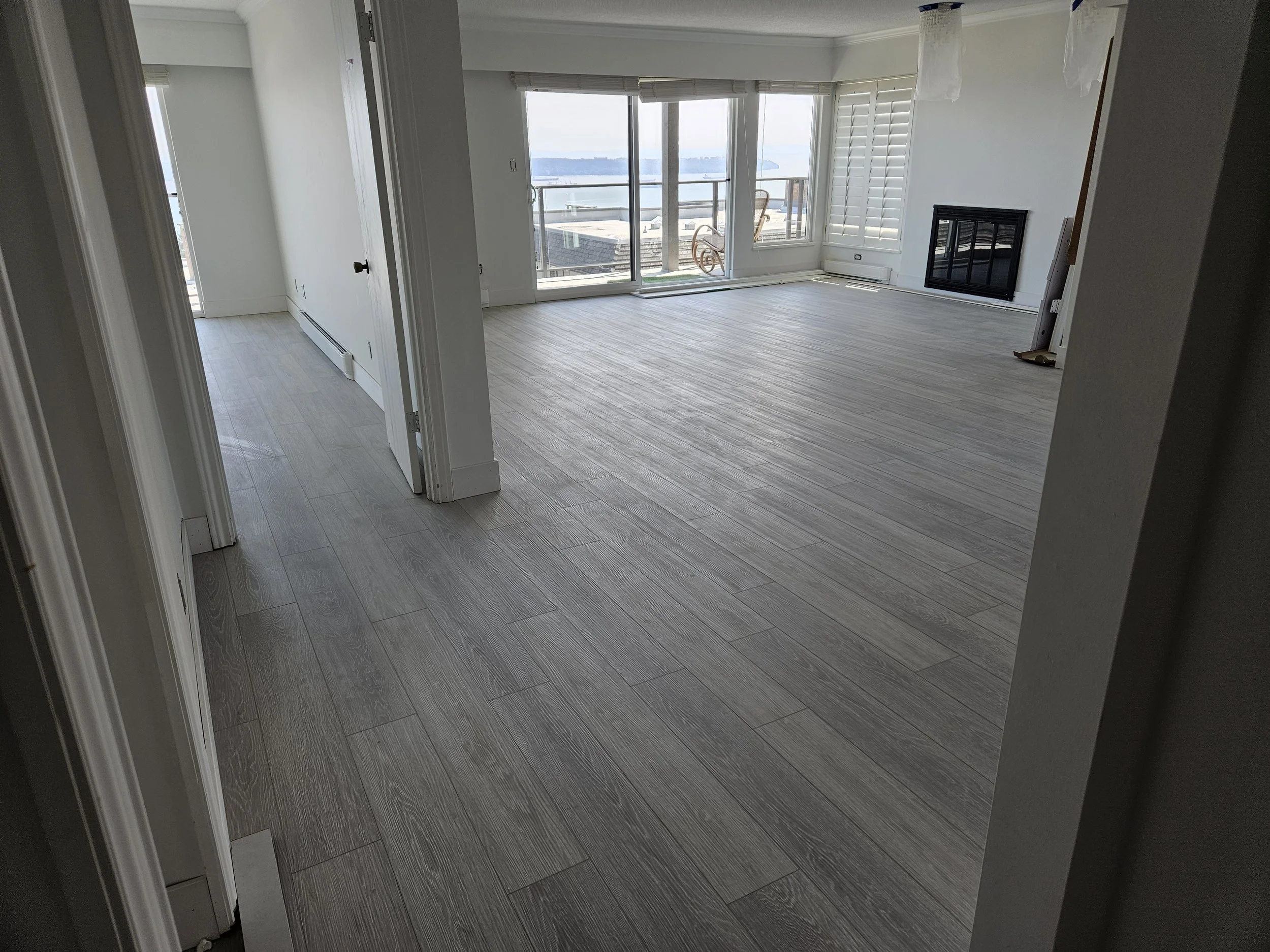 Empty living room with large sliding glass doors leading to a balcony, a fireplace, and white walls. The room has light-colored wood-look flooring and window shutters.
