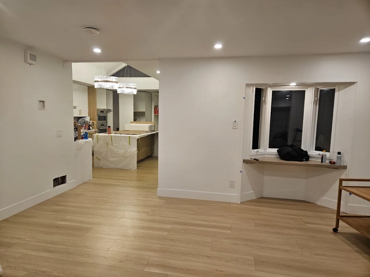 Living room and kitchen area under renovation, with tools and supplies on counters and window ledge, and wooden flooring.