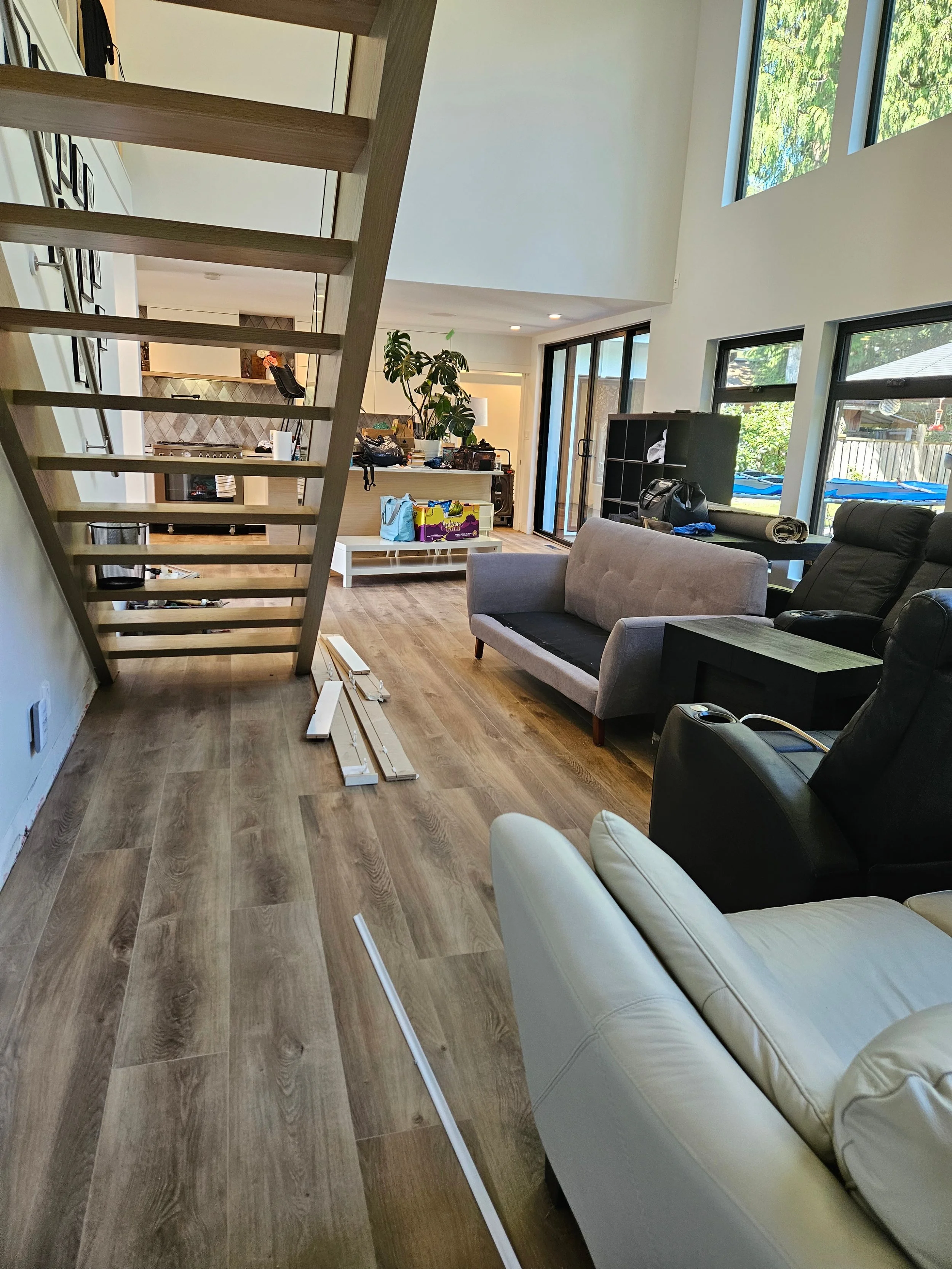 Interior of a modern living room with wooden flooring, large windows, a staircase, and various furniture and items scattered around.