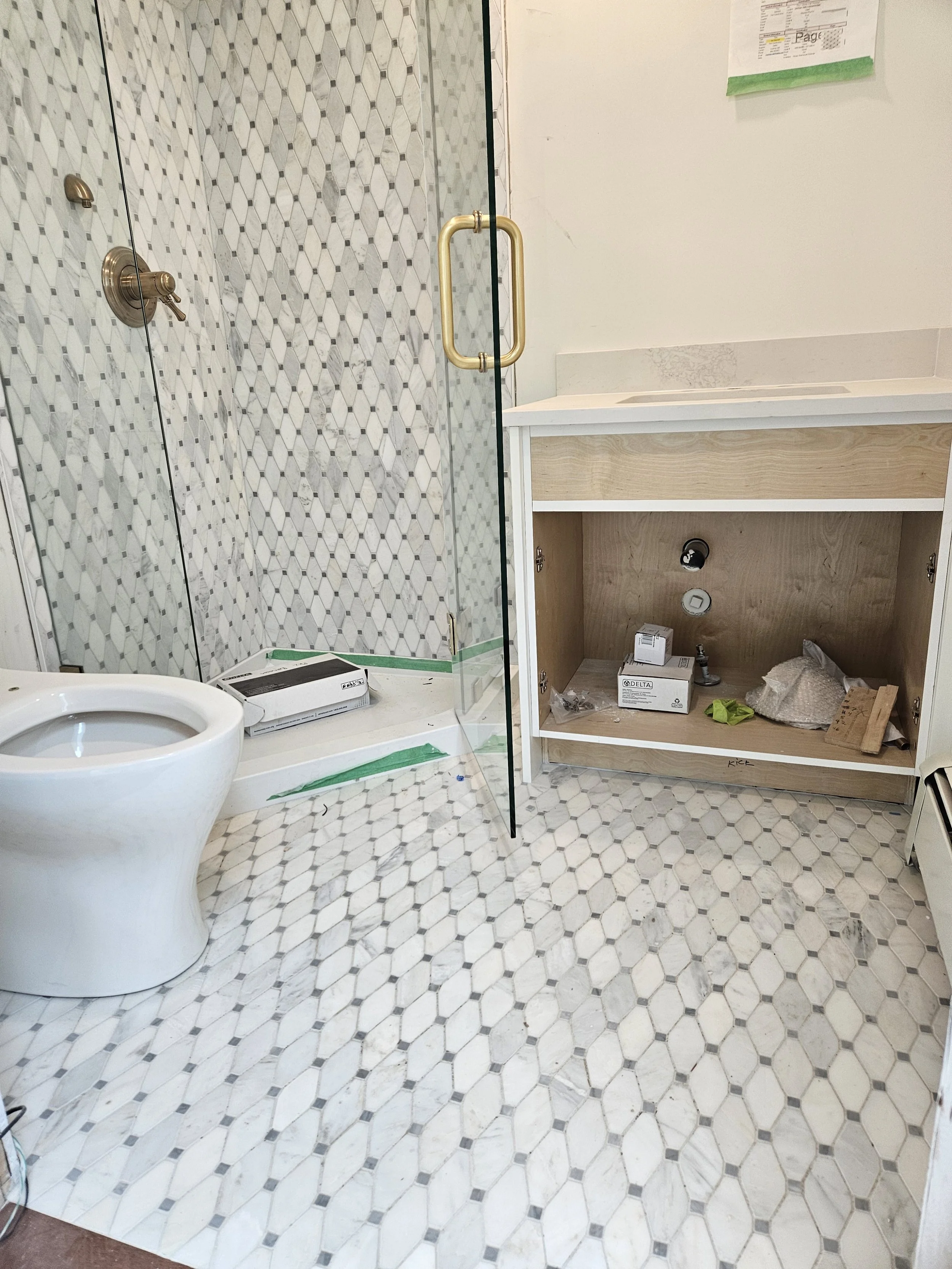 Bathroom with white and gray hexagonal tile floor, a glass shower enclosure with brass handles and fixtures, an empty vanity cabinet beneath a white countertop with an integrated sink, and a mirror on the wall.
