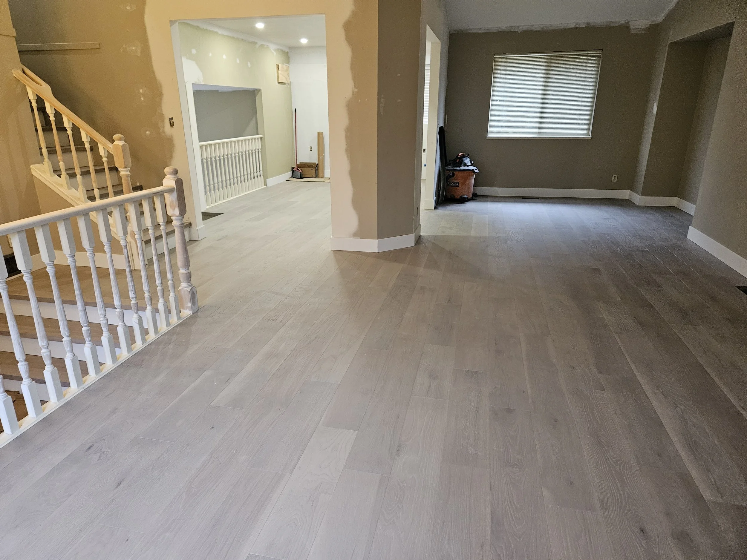 Living room with light-colored wooden flooring, beige and gray walls, a window with blinds, and some construction tools and materials in the background.