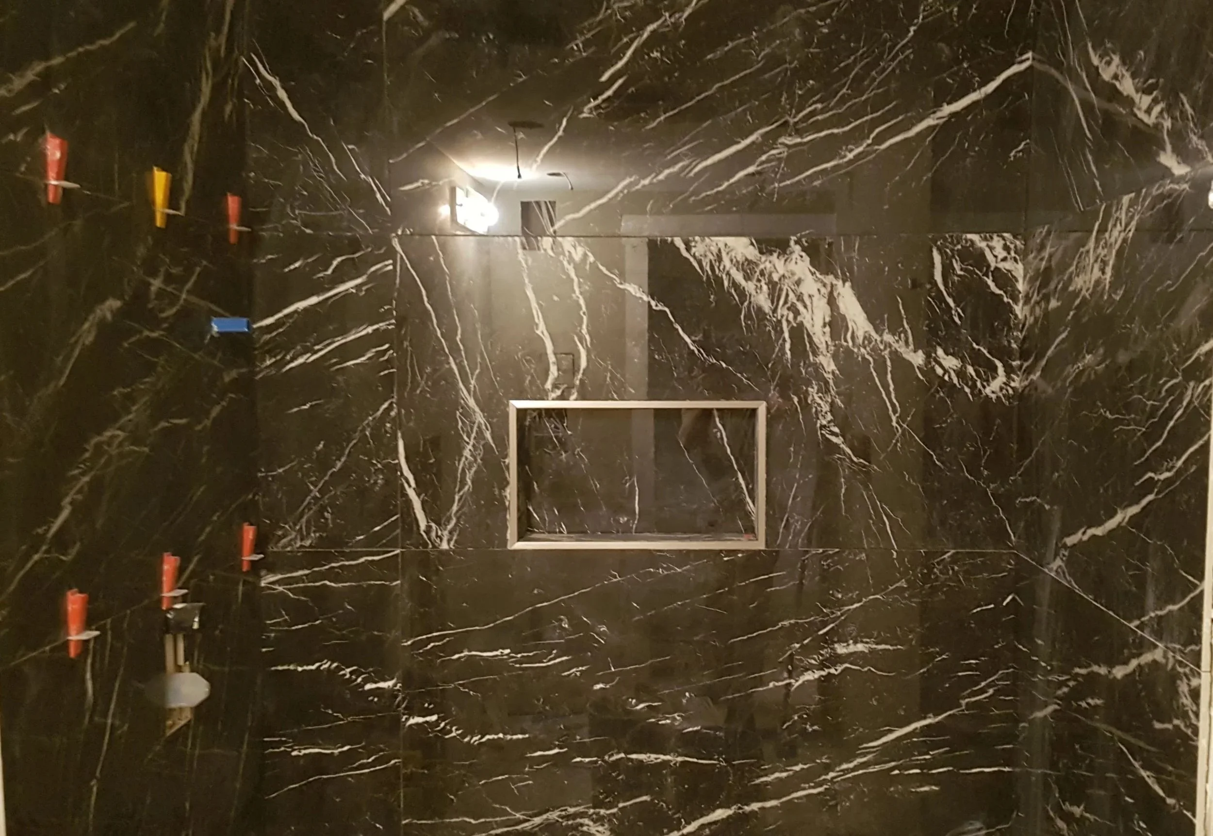 Black marble shower wall with white veining, a recessed niche, and shower fixtures covered in red, yellow, blue, and gray caps.
