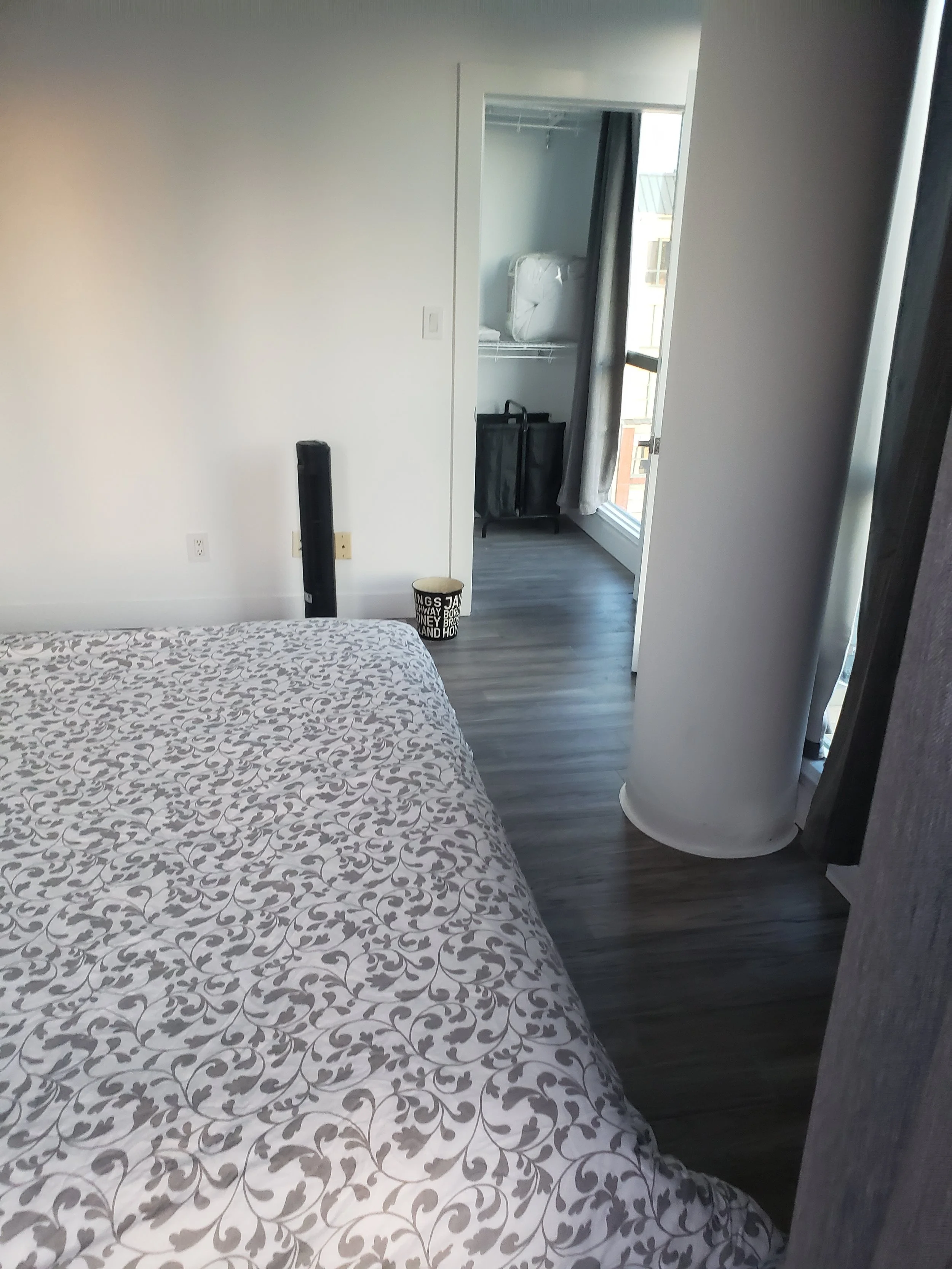 A bedroom with a bed featuring a white and gray floral pattern. Near the wall is a black tower fan and a small trash can. At the end of the room, there's a door leading to a balcony with a view of nearby buildings. Closet with a black trolley and a w