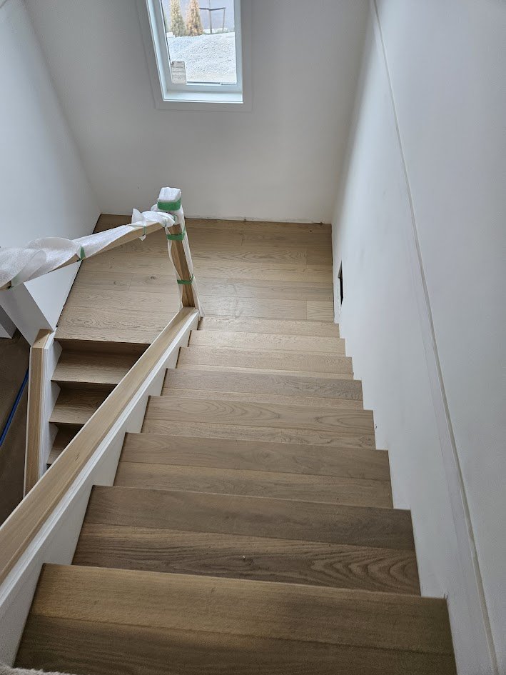 Wooden staircase under construction with protective wrapping on the handrail, leading down to a small landing with a window showing a gravel yard outside.