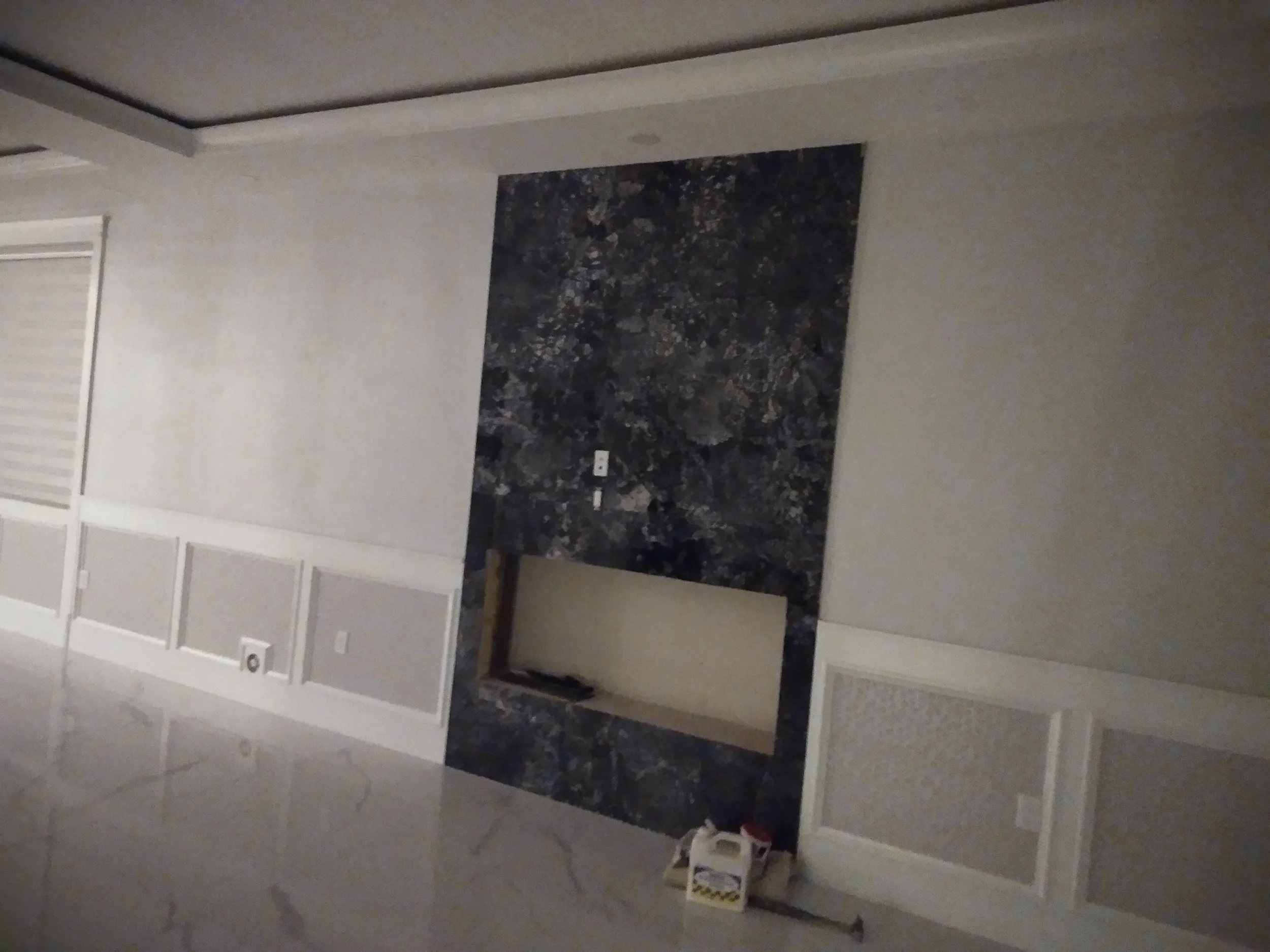 Interior of a room with a dark marble fireplace, partially built-in, with some tools and materials nearby, cream-colored walls with decorative paneling, and a window with closed blinds.