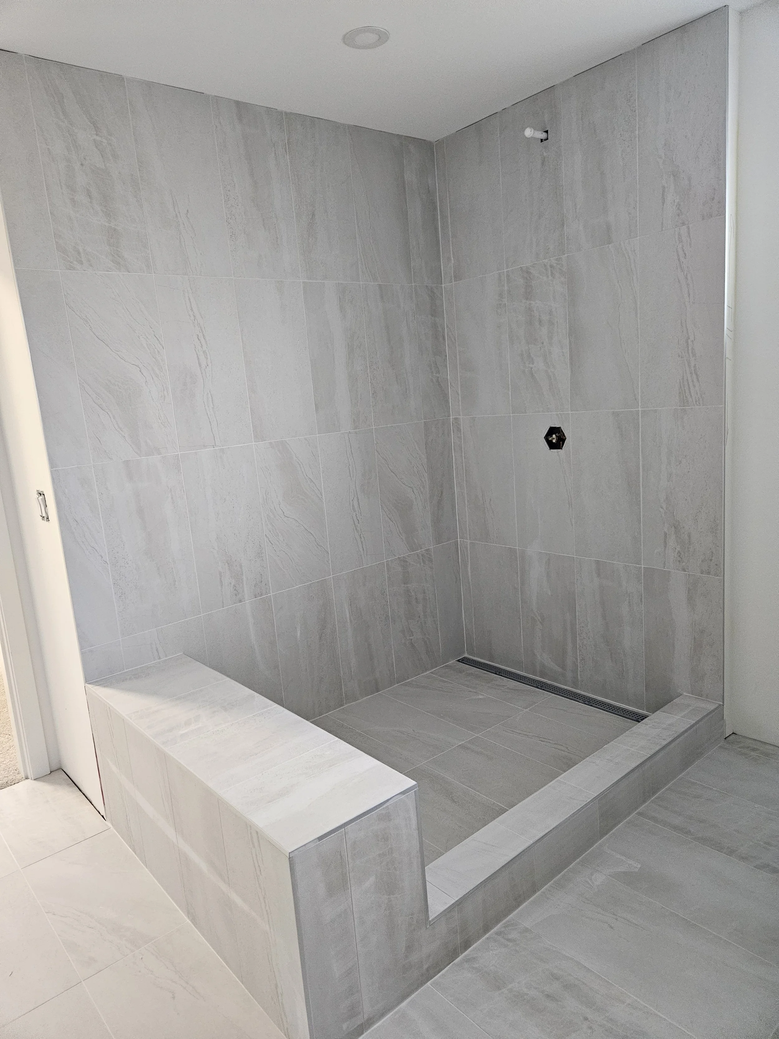A modern walk-in shower with gray tiled walls and floor, a built-in bench, and a linear drain, with a small window on the right side.