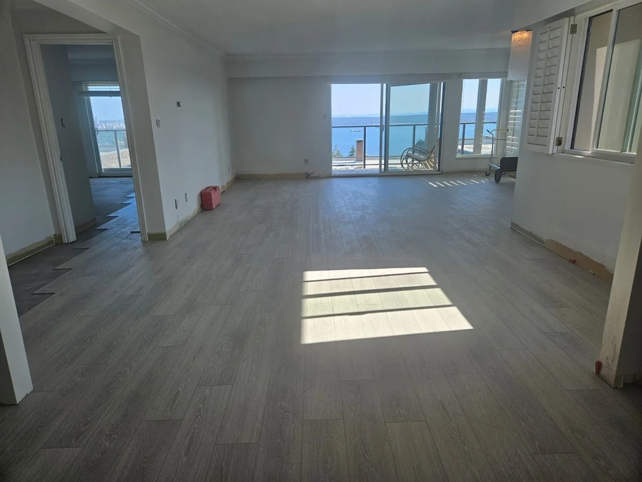 Empty living room with large windows and sliding doors showing a view of water and sky, hardwood floors, and some construction or renovation materials.