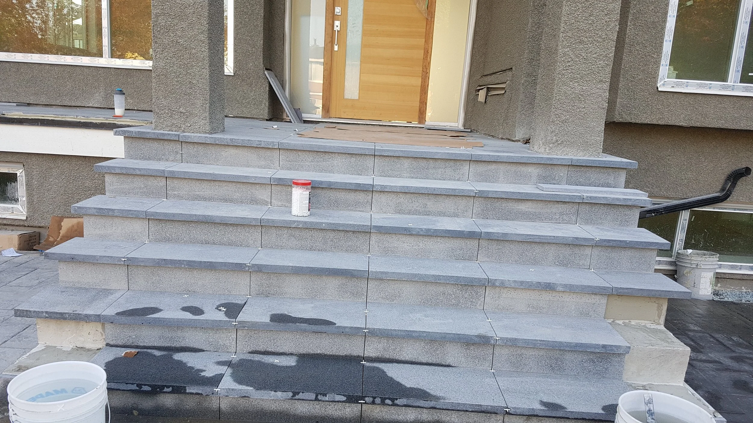 Construction site showing gray tiled stairs leading to a wooden front door of a house under construction, with tools and building materials around.