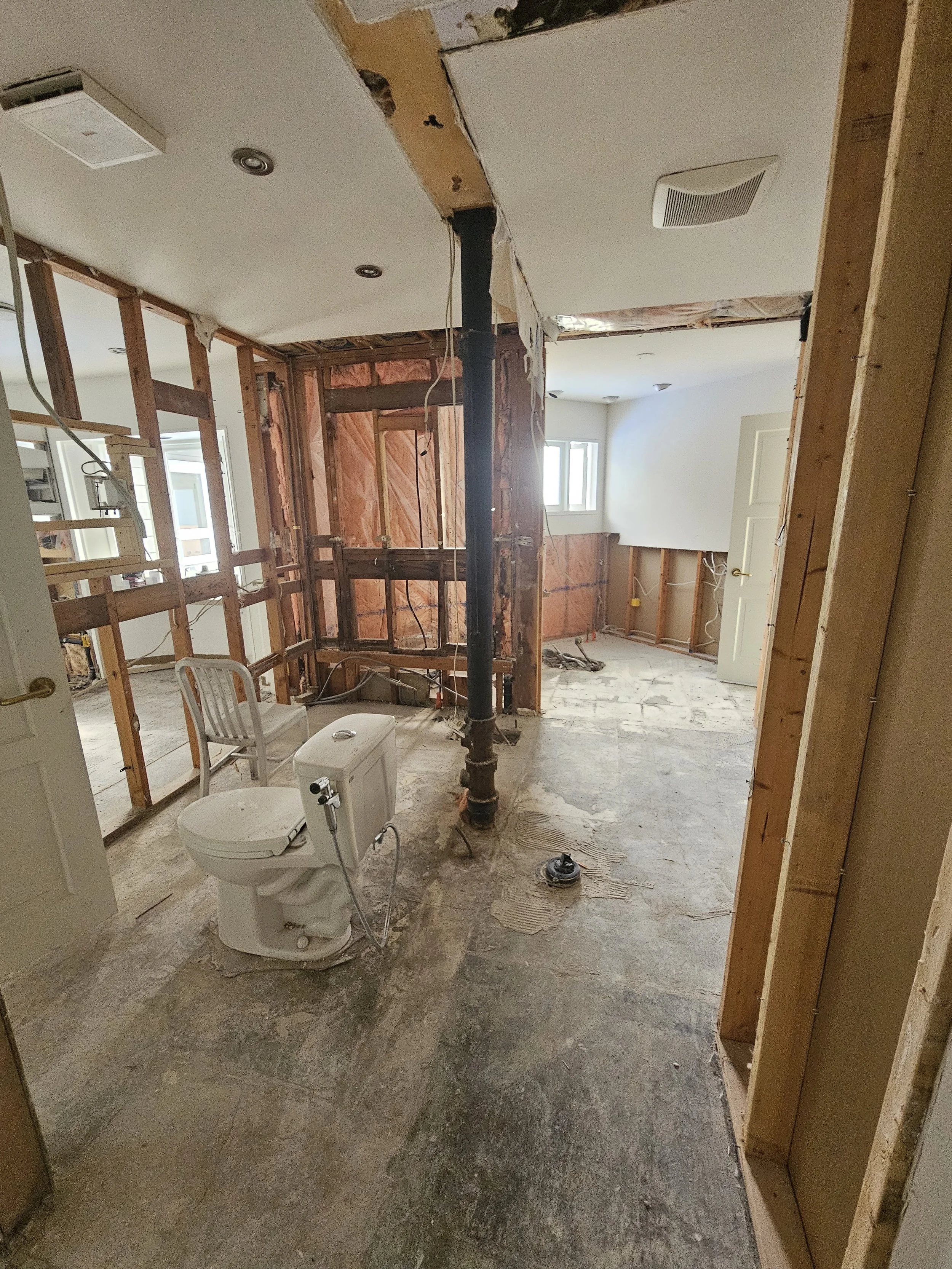 Bathroom under renovation with exposed drywall, pipes, and electrical wiring, with a toilet, chair, and construction materials visible.