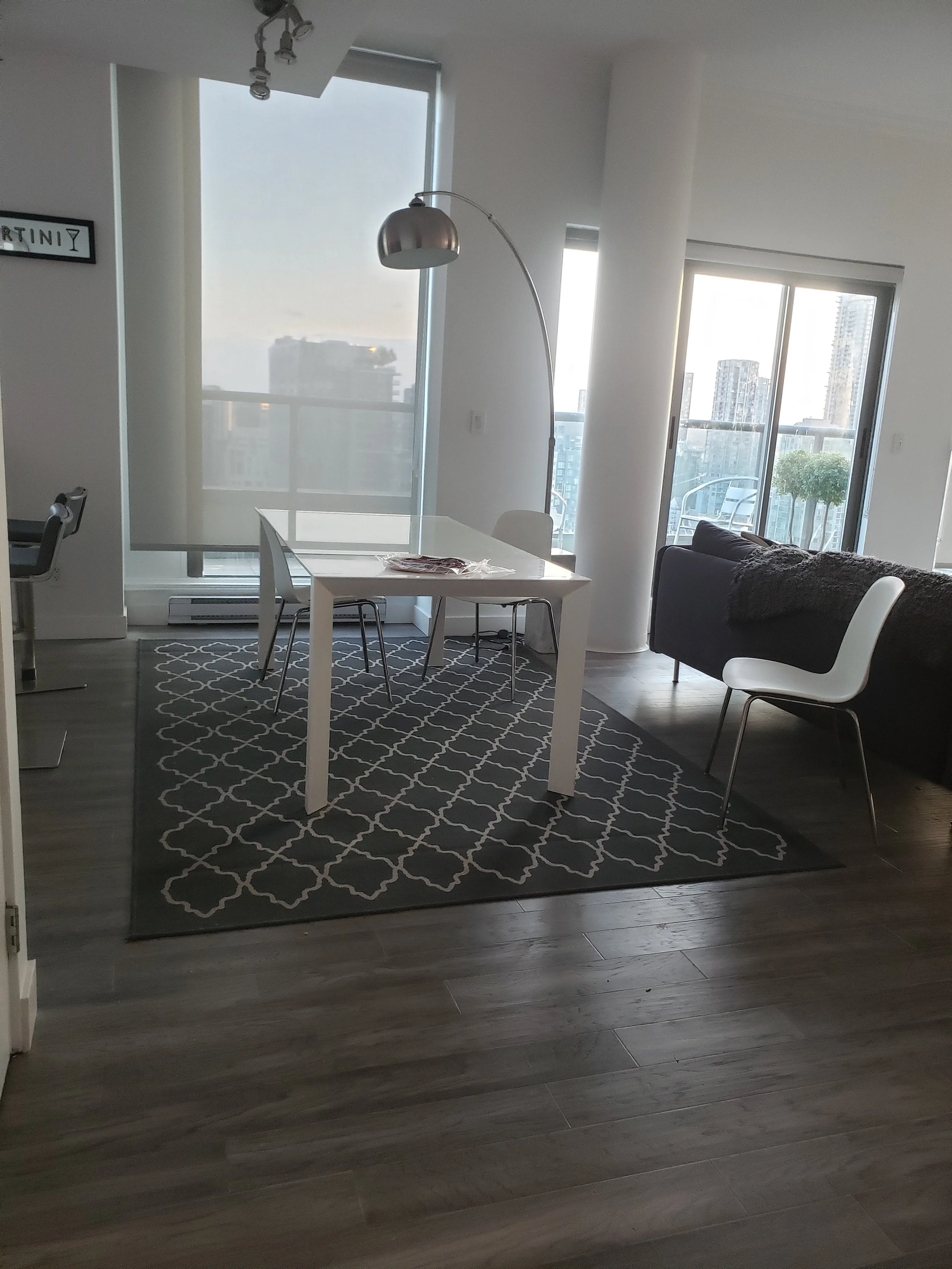 Modern dining area with a white table, four white chairs, and a geometric-patterned black and white rug. Large windows let in natural light, with city buildings visible outside. A black sofa with a throw blanket is in the background, and a tall, arched floor lamp is beside the table.