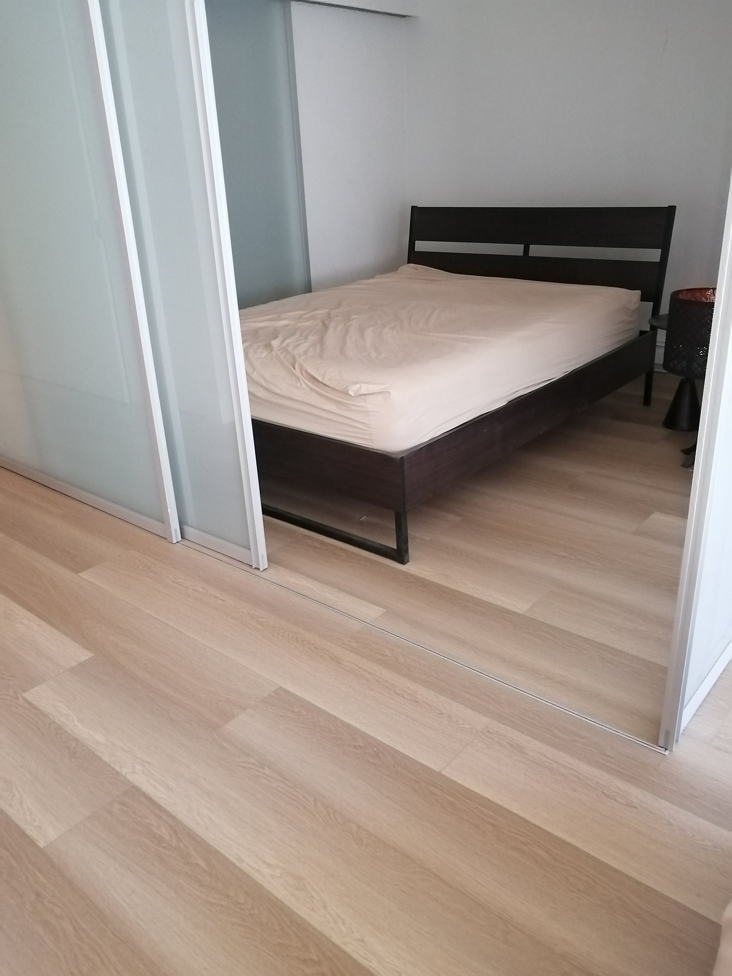 A bedroom with a black wooden bed frame and an unmade beige mattress, a small black nightstand with a black basket, and a sliding frosted glass door closet, on a light wood floor.