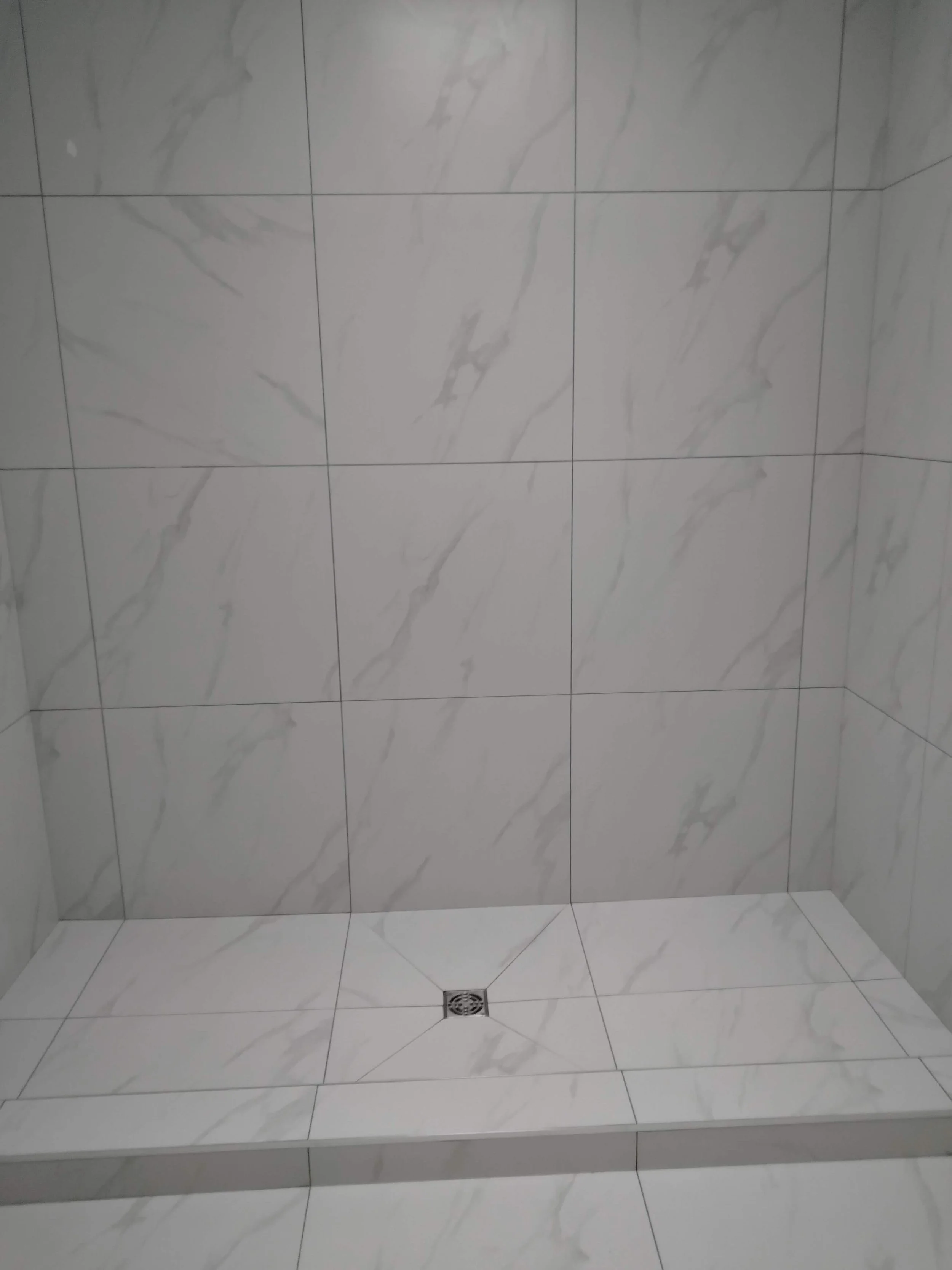 Empty tiled shower with marble-patterned tiles, a drain at the center of the floor, and tiled walls with a corner visible.
