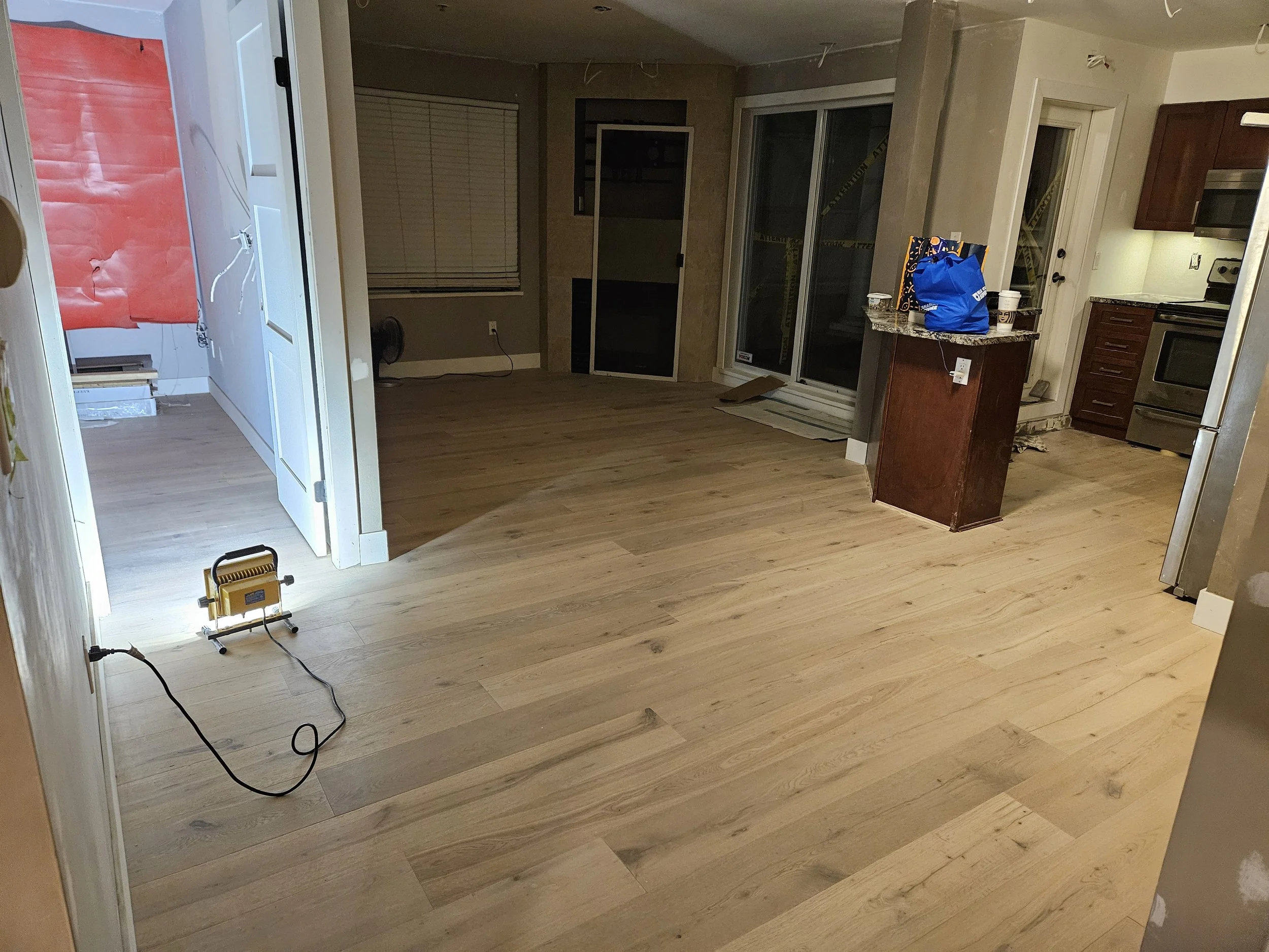 Empty kitchen and living area with light wood flooring, partially installed, with a portable work light on the floor, and a blue bag and two cups on a small kitchen counter.