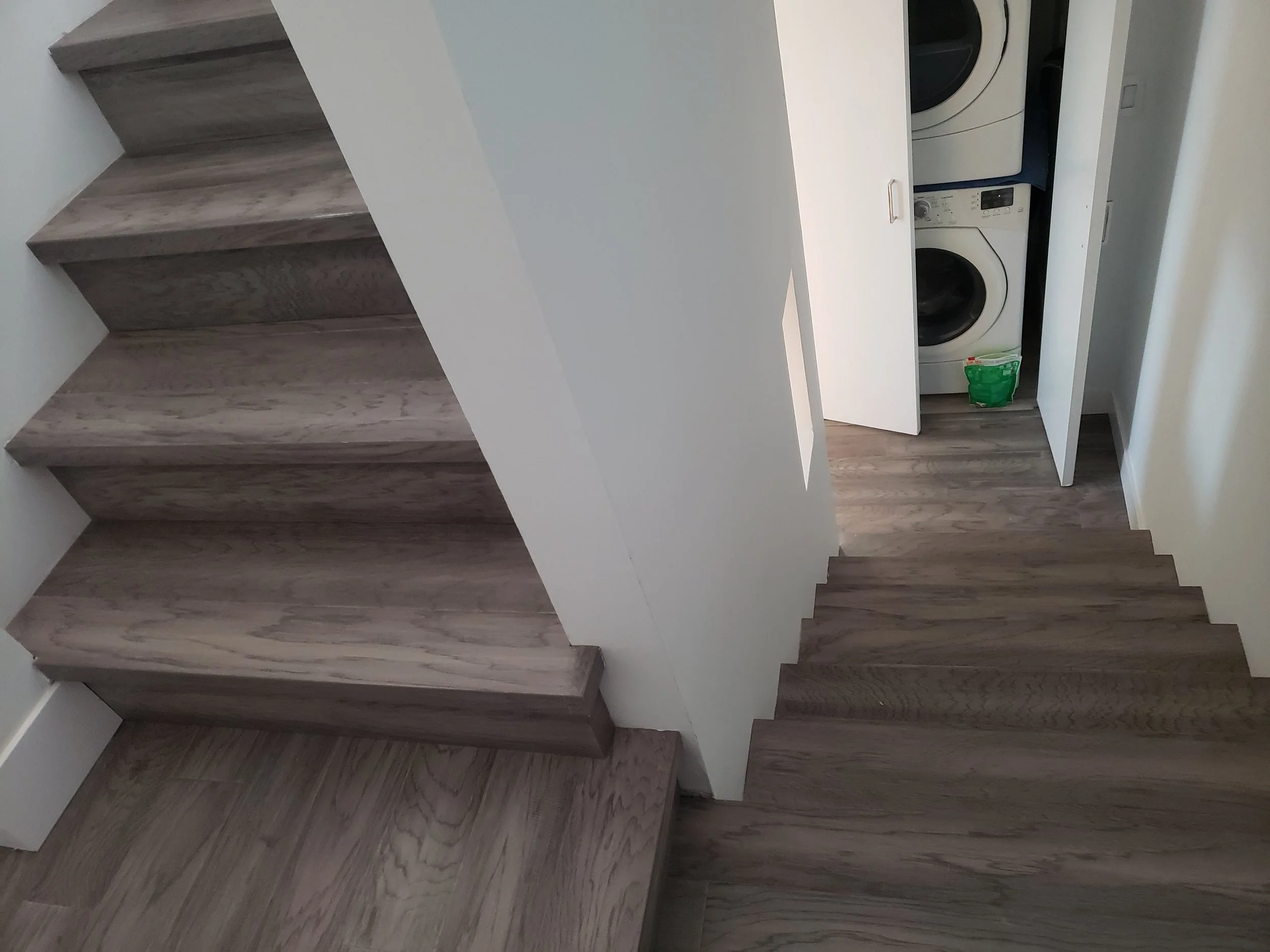 Interior view of a staircase with wooden steps leading down to a small laundry area with a washing machine and a bag of laundry detergent inside a closet.