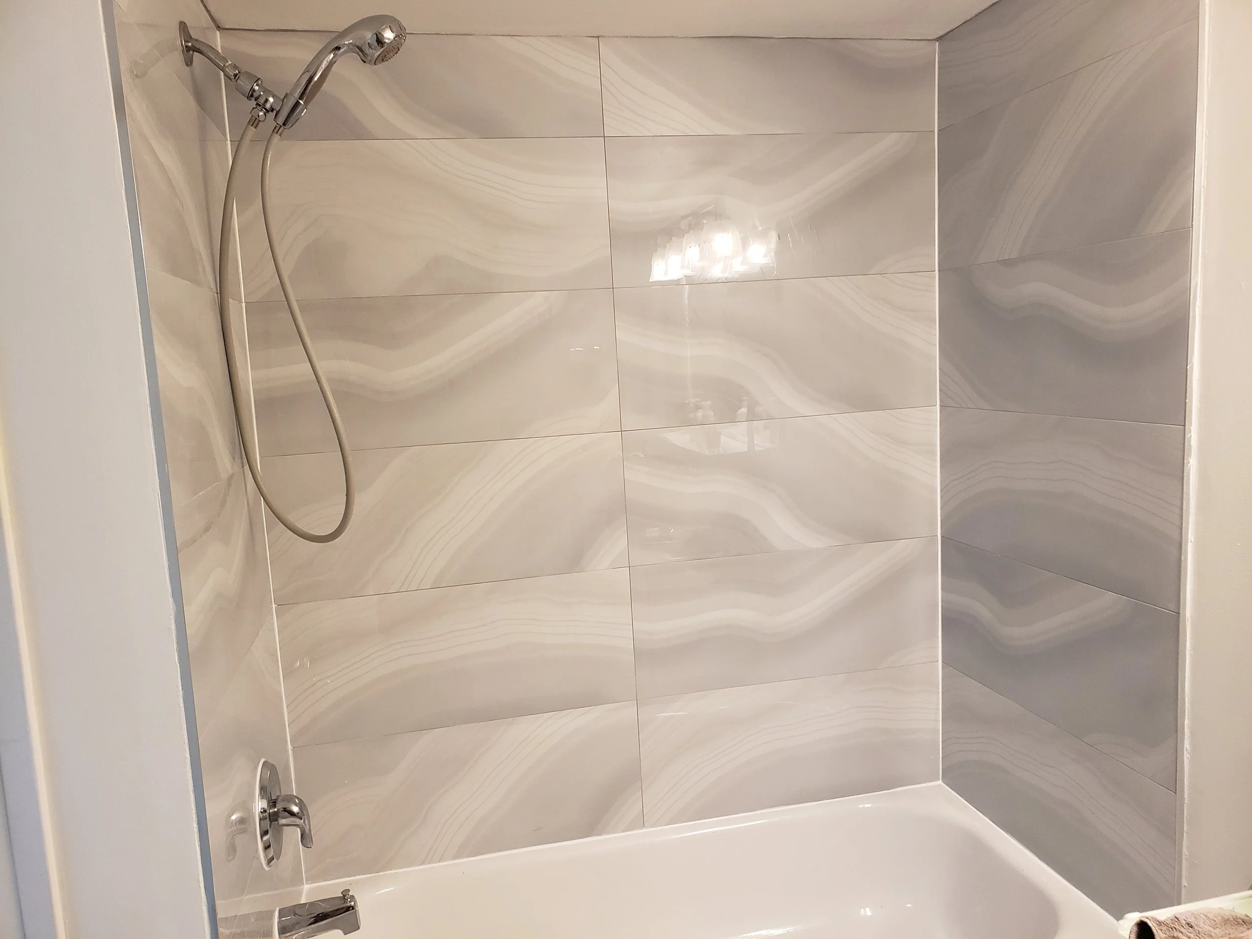 Bathroom shower area with beige marble-patterned tiles on the wall, a chrome showerhead with a flexible hose, a faucet, and a white bathtub with a towel in the corner.