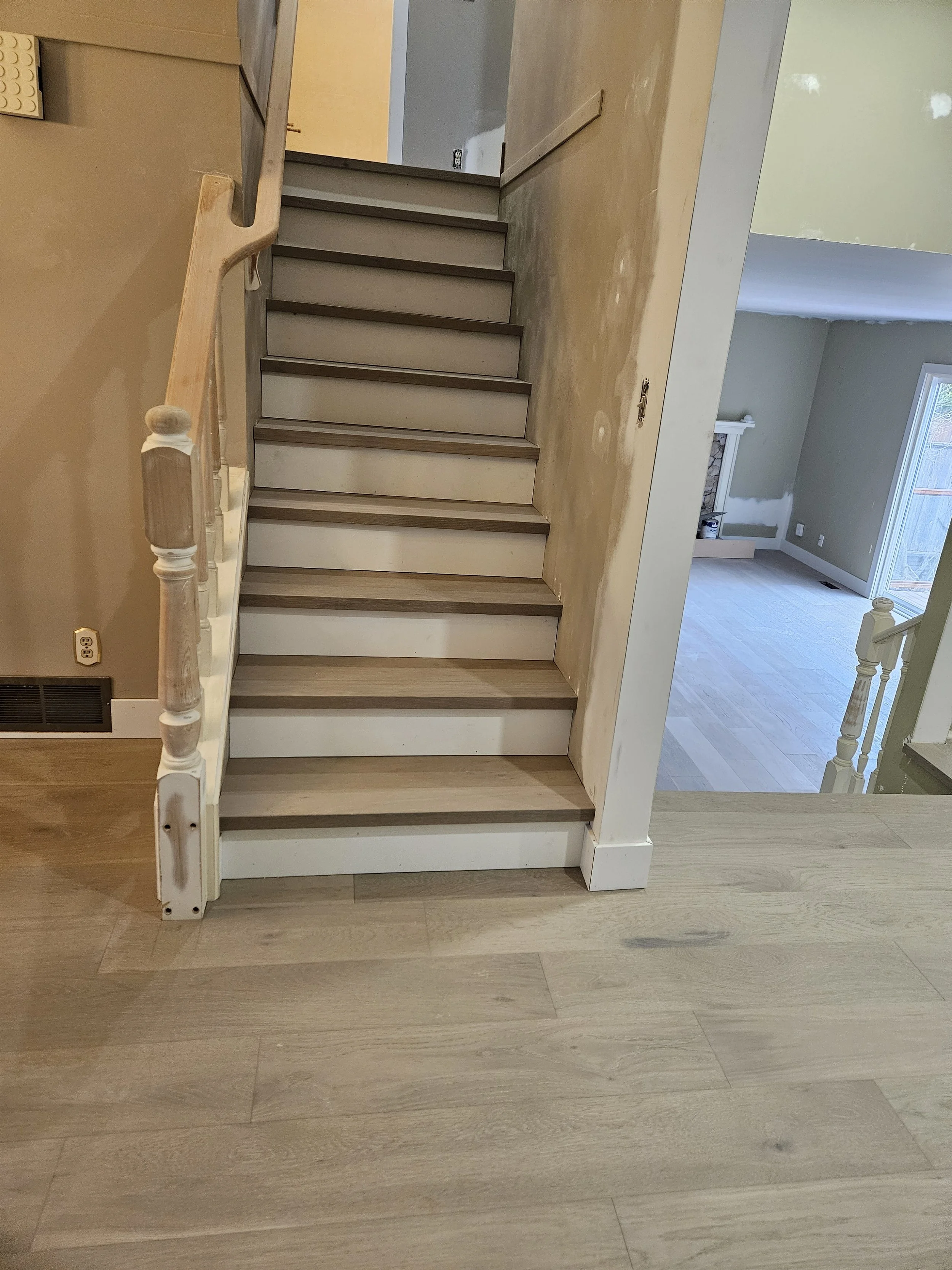 Interior view of a wooden staircase under construction with unfinished walls and flooring, and a living room with large sliding glass door in the background.