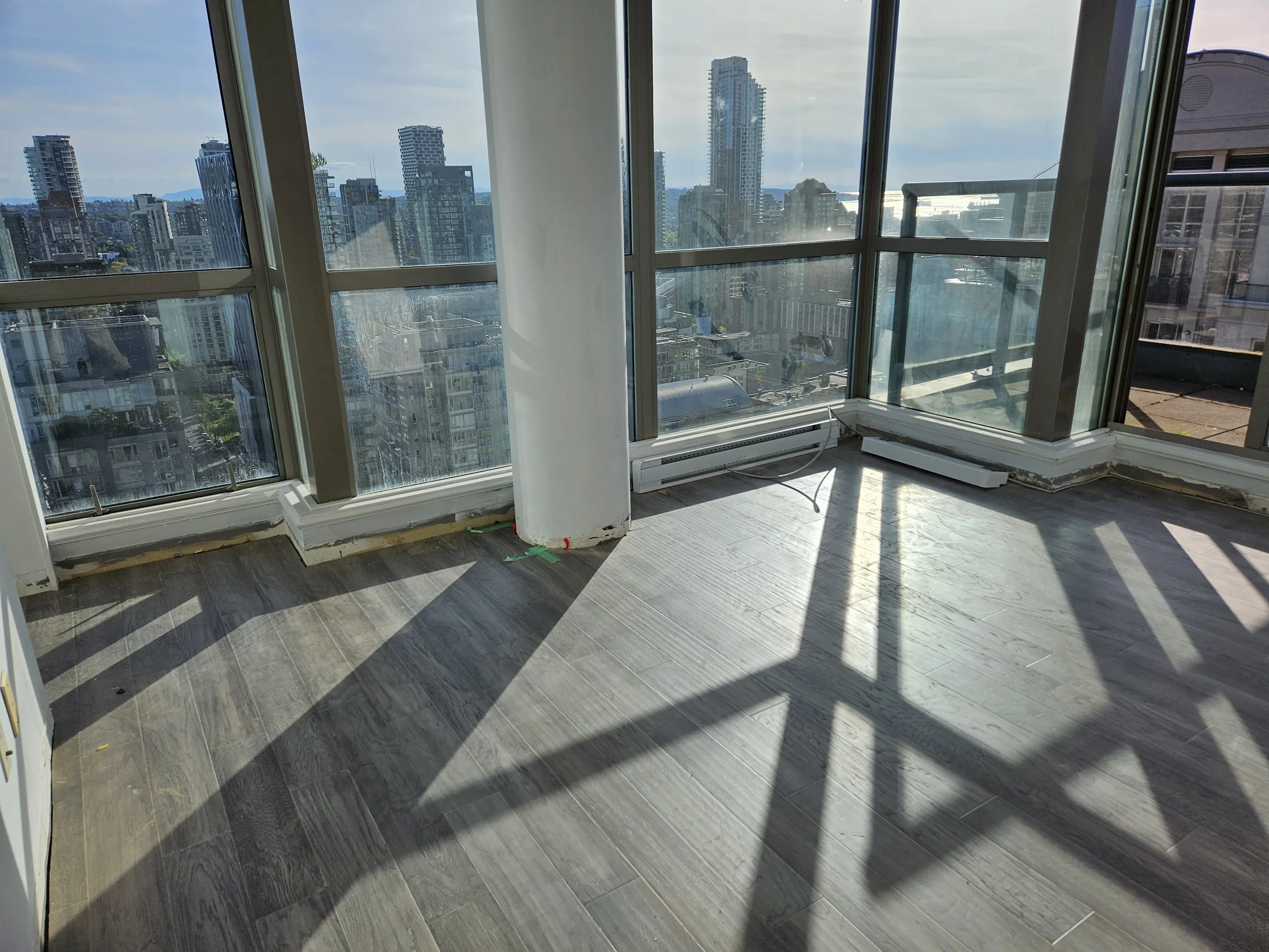 Empty room with large floor-to-ceiling windows, city skyline view, hardwood floors, and shadows cast by window frames
