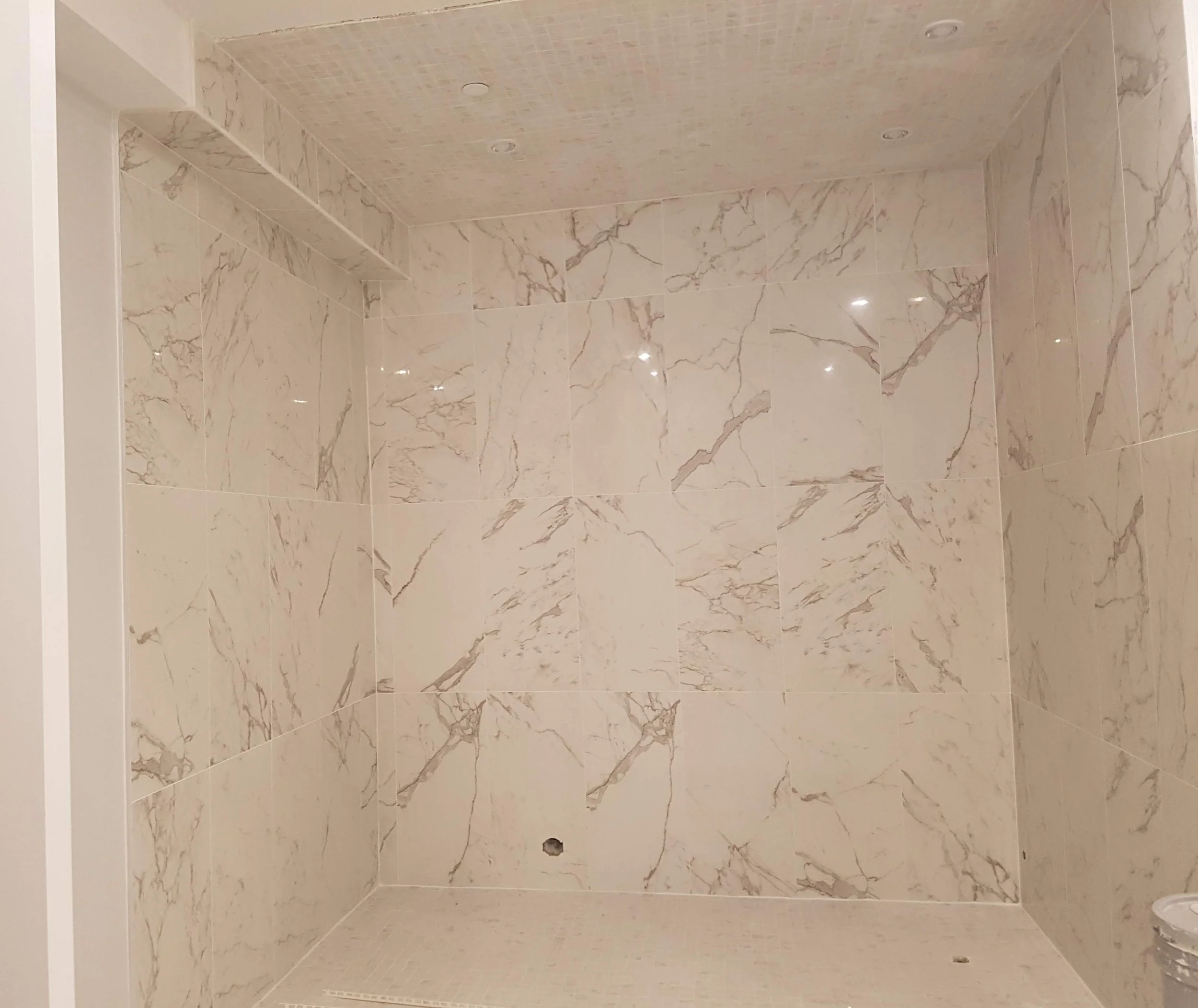 Empty walk-in shower with beige marble tiles on walls and ceiling, and small beige tiles on the shower floor. A drain is visible on the floor, and a small hole near the bottom of the wall.
