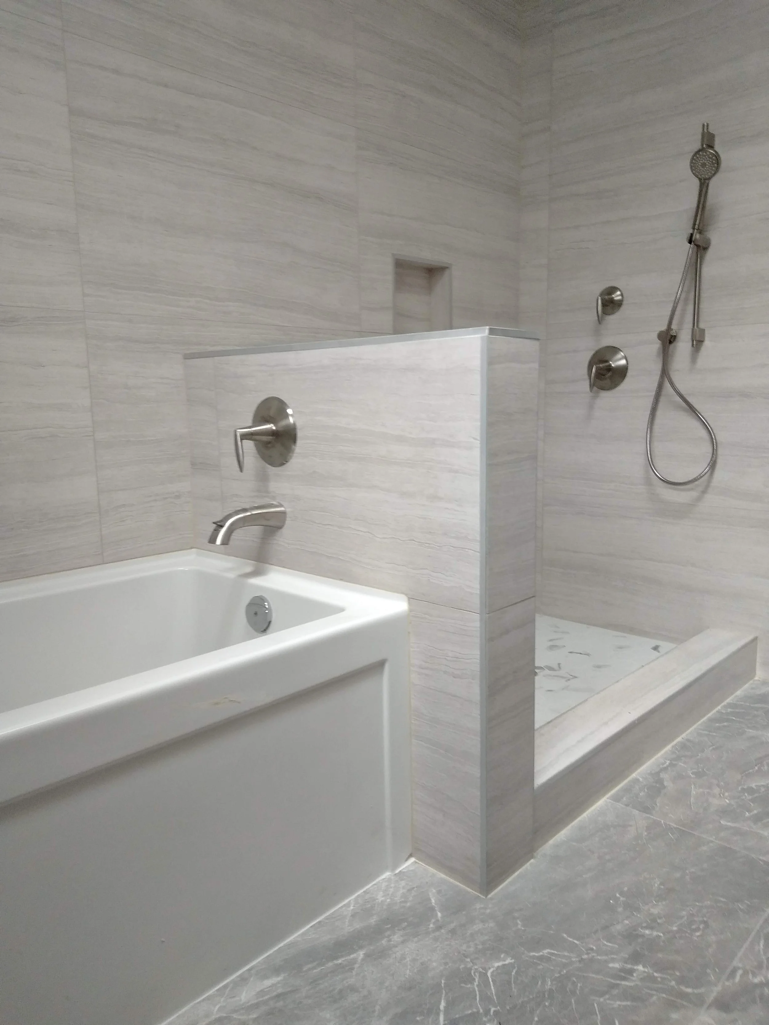 Modern bathroom with a white bathtub, brushed nickel fixtures, and a walk-in shower with a handheld showerhead, beige tiled walls, and gray marble floor.