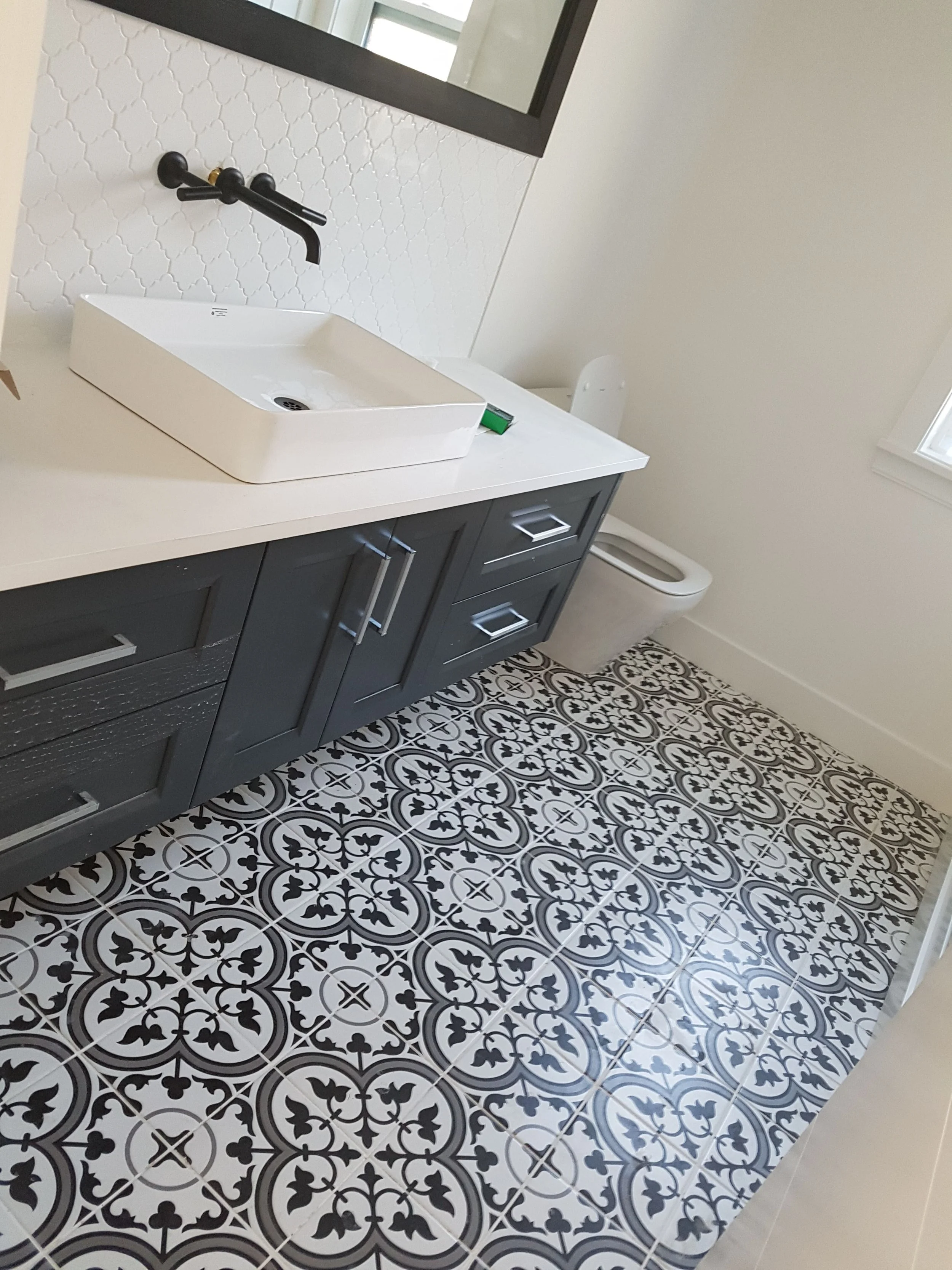 A modern bathroom with black and white patterned tile floor, dark gray vanity with silver handles, white vessel sink, black wall-mounted faucet, and a white toilet. There is a large mirror above the sink and a window on the right.