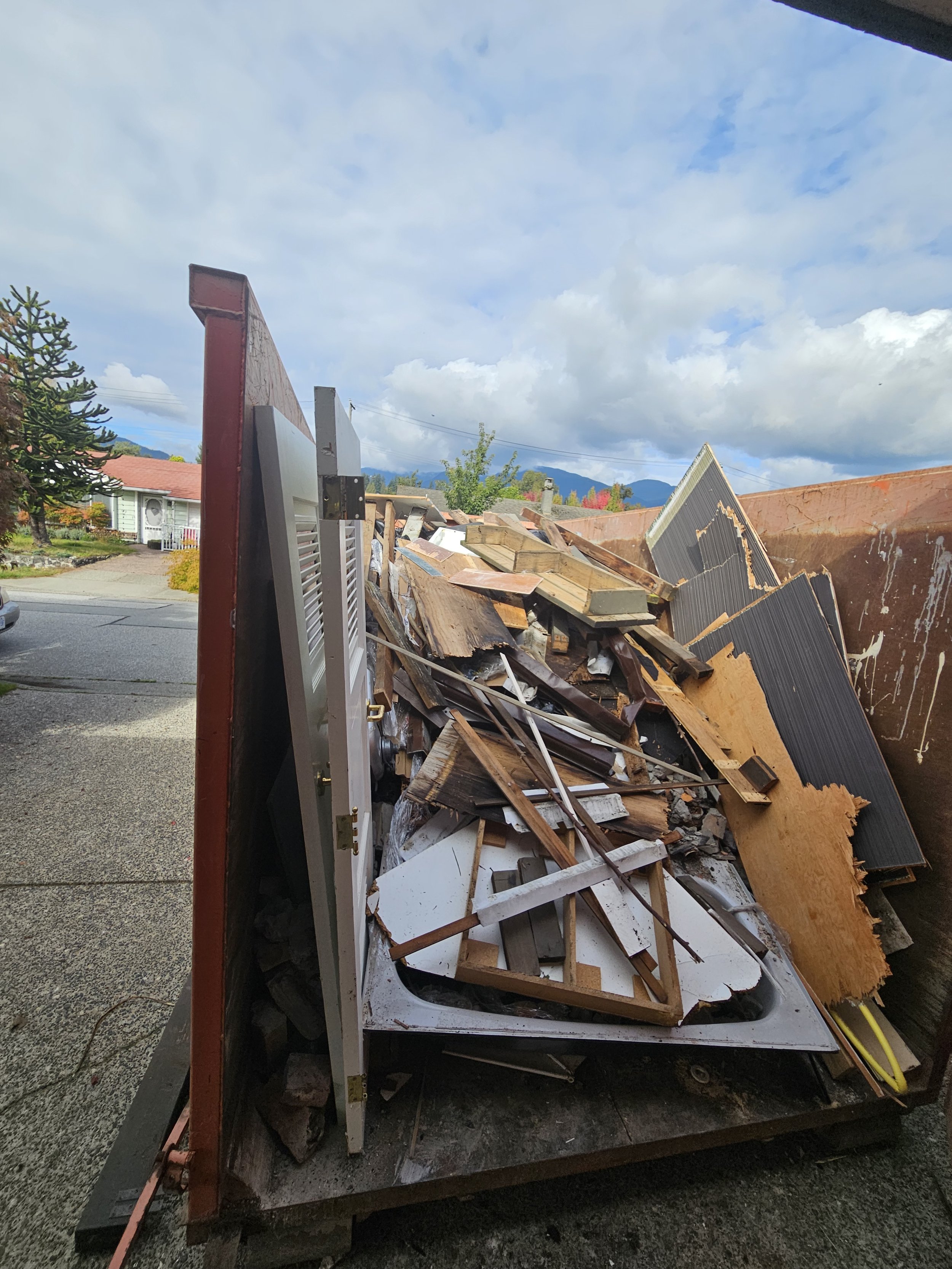 A trailer filled with debris, including wooden planks, broken furniture, and other construction or demolition materials, parked on a residential street with houses, trees, and mountains in the background.