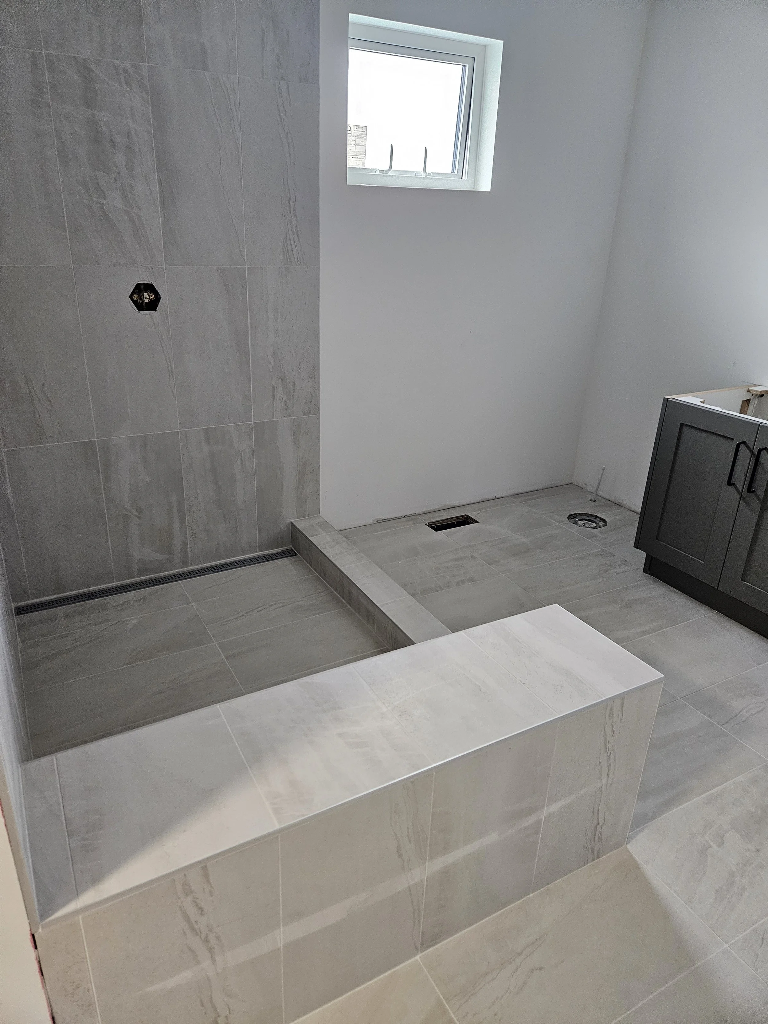 Bathroom under construction with tiled shower area, window, and a gray vanity cabinet.