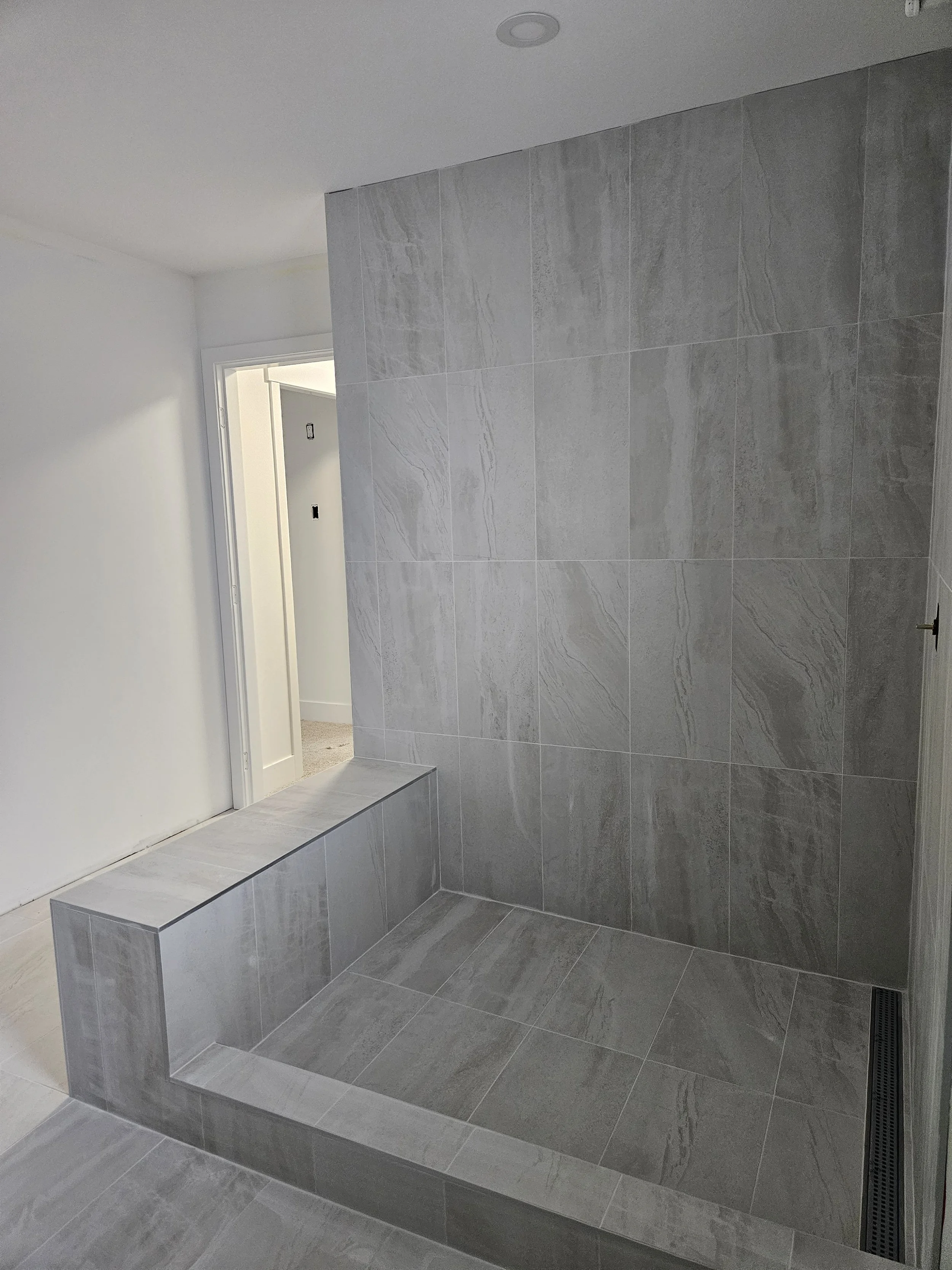 A modern tiled walk-in shower with grey stone tiles, a small bench, and a ceiling light, adjacent to an open doorway leading to a room with carpeted flooring.