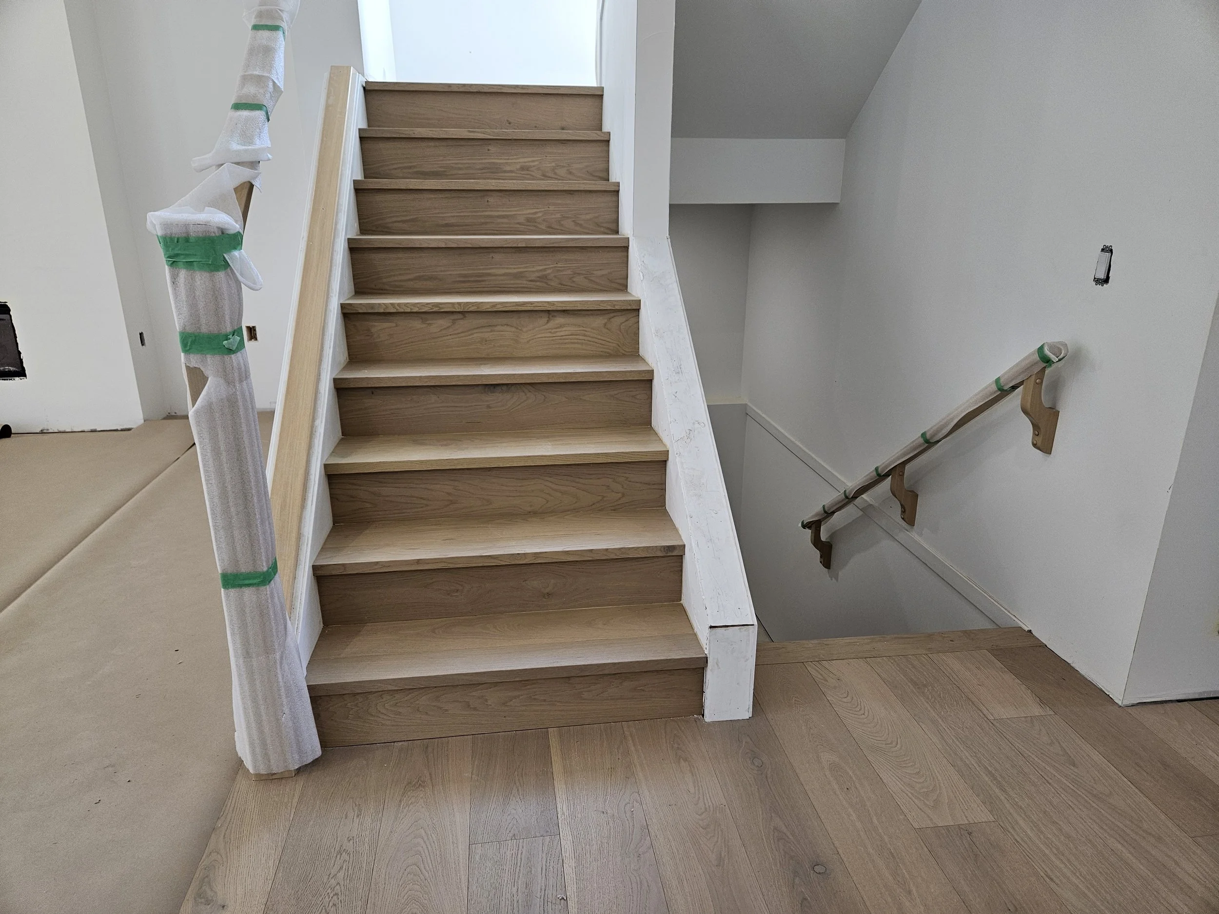 Wooden staircase with a white wall and a handrail wrapped in protective covering, likely in a home under renovation.