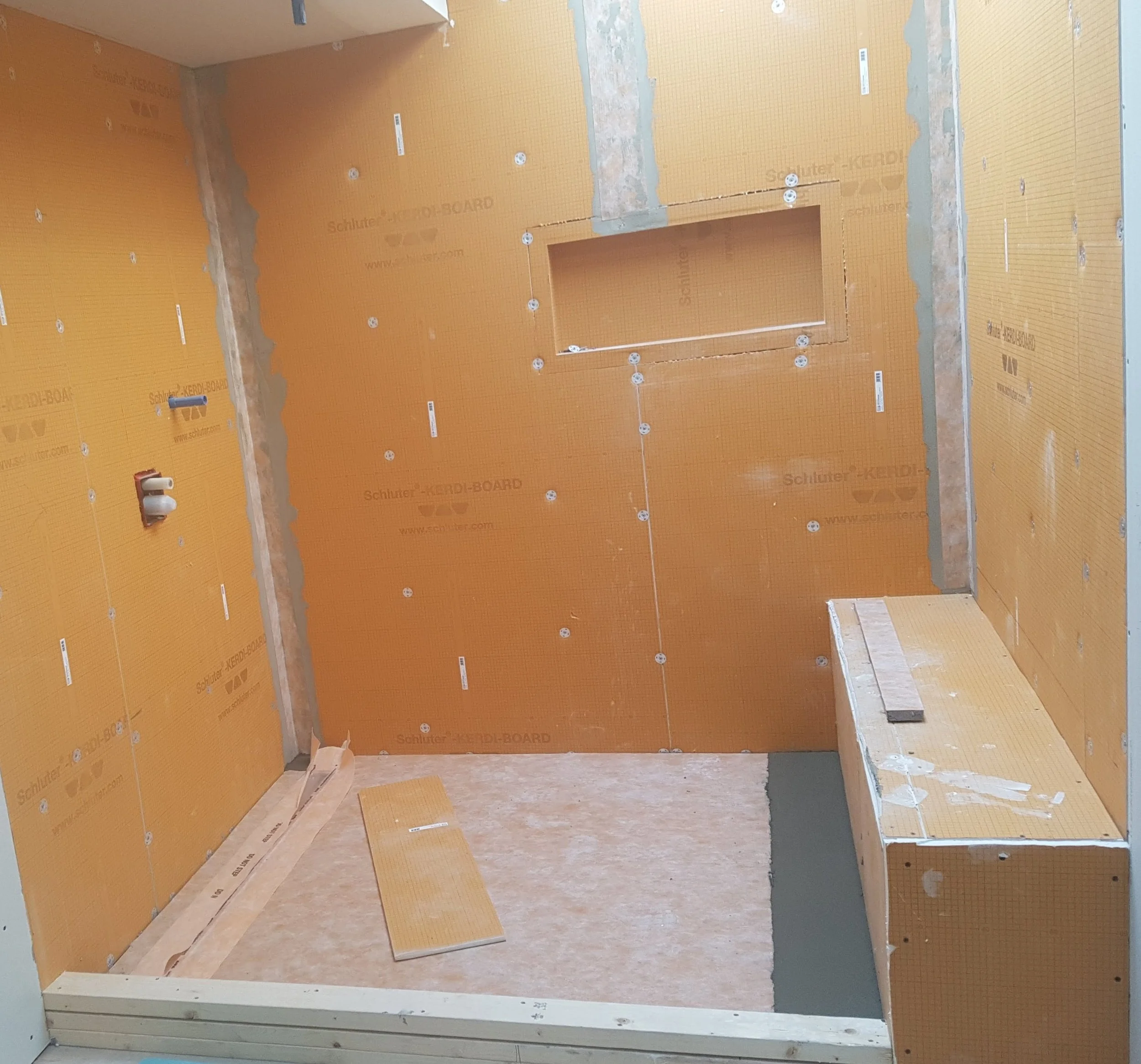 A partially finished bathroom with orange waterproof wall panels, a built-in niche, unfinished floor, and a wooden bench.